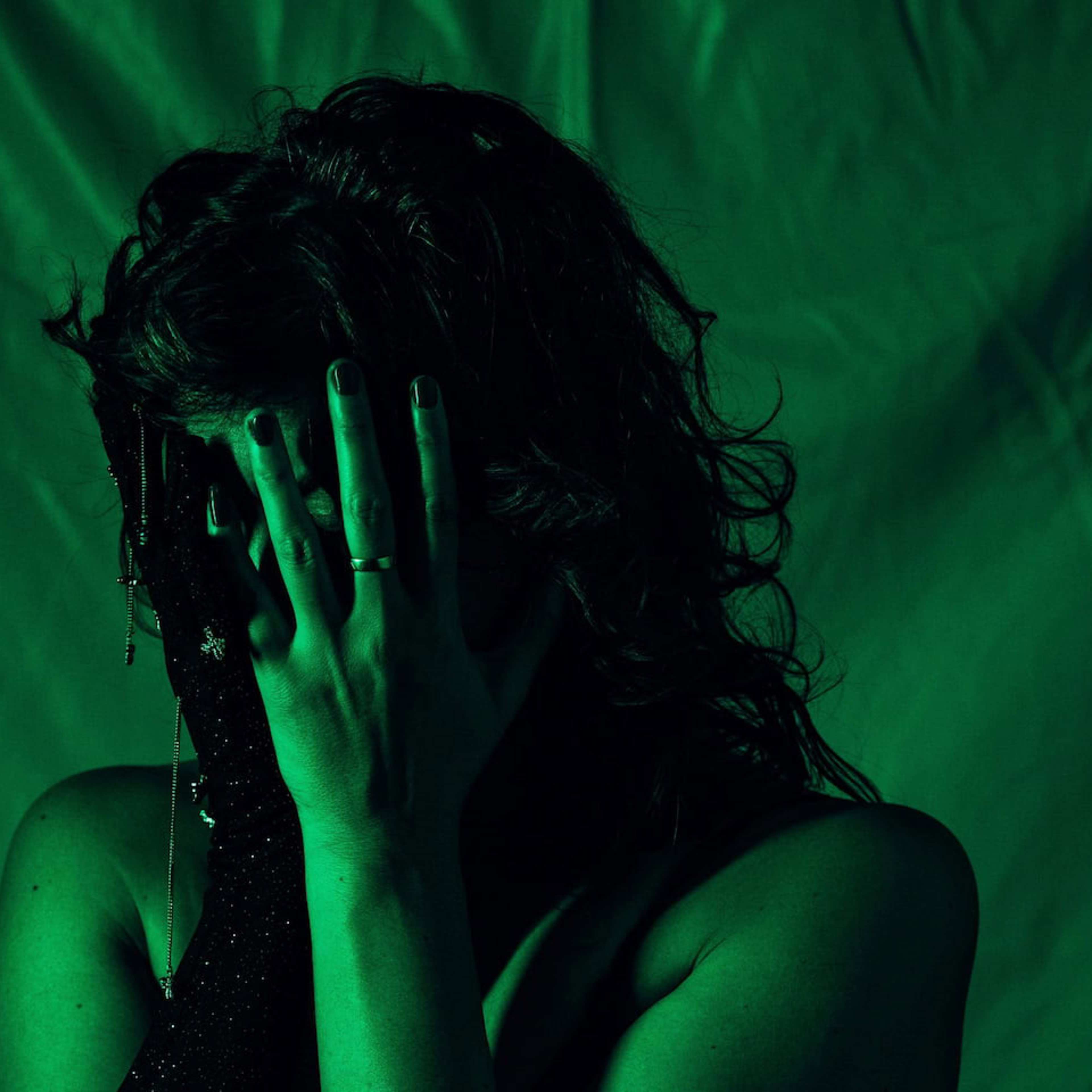 A person under green lighting, with long dark curly hair, covering their face with their hands, wearing on black, sparkly glove.