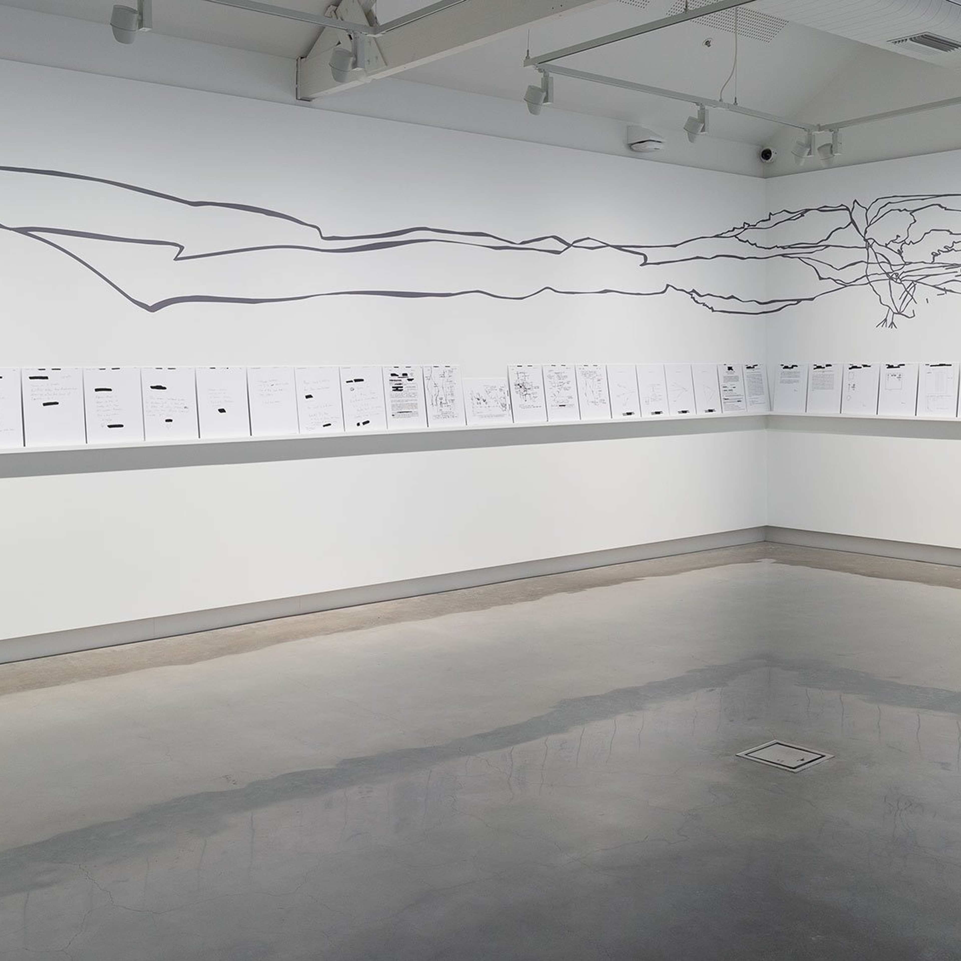 A minimalist art gallery display features documents lined up on a white shelf along two walls, beneath abstract black line art resembling mountain ranges drawn directly on the walls. The floor is polished concrete.