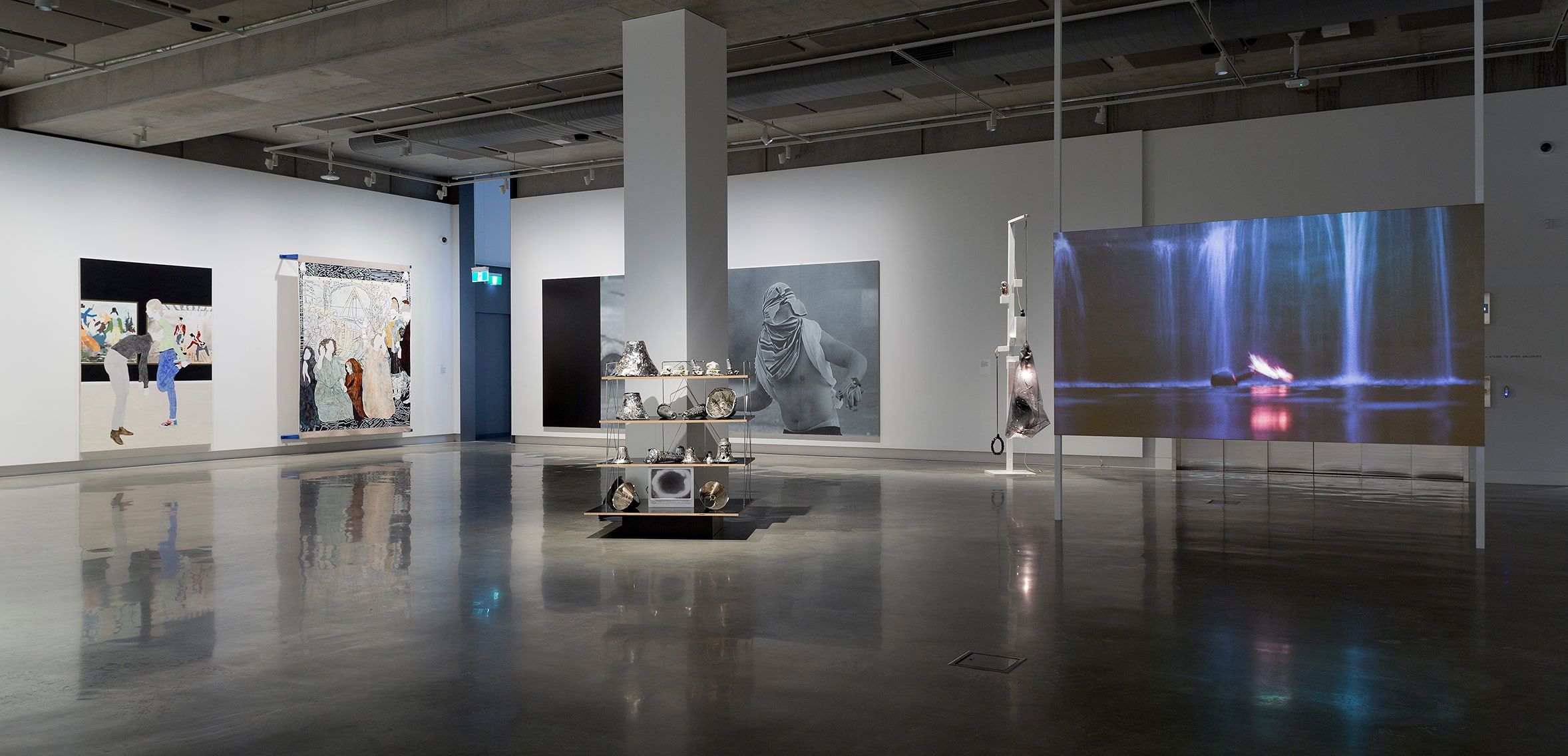 A modern art gallery with polished concrete floors, large paintings on white walls, a sculpture display in the center, and a large screen showing a video of a person floating in water. The space is well-lit and spacious.