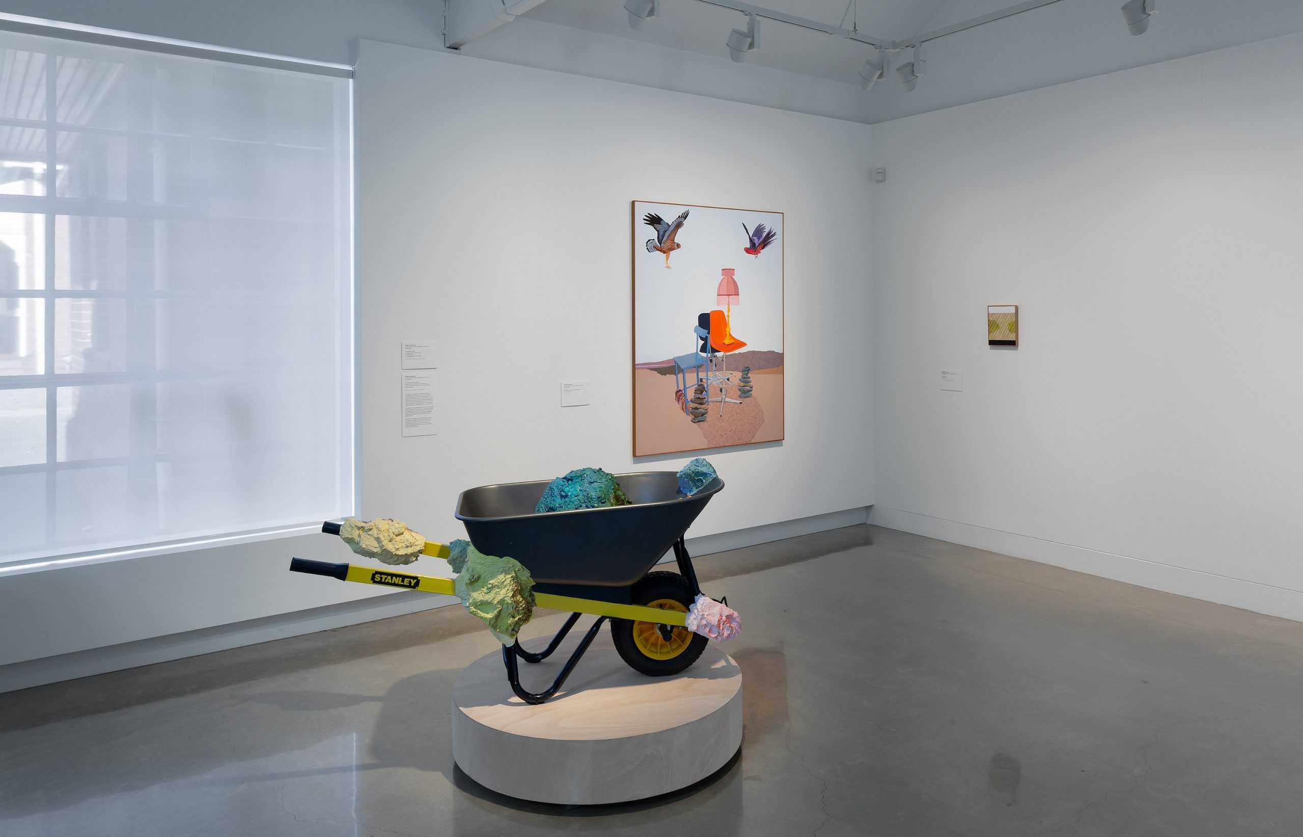 A modern art gallery with a wheelbarrow sculpture filled with colorful rocks on a pedestal; behind it, a framed artwork of birds, a lighthouse, and abstract shapes hangs on the white wall.