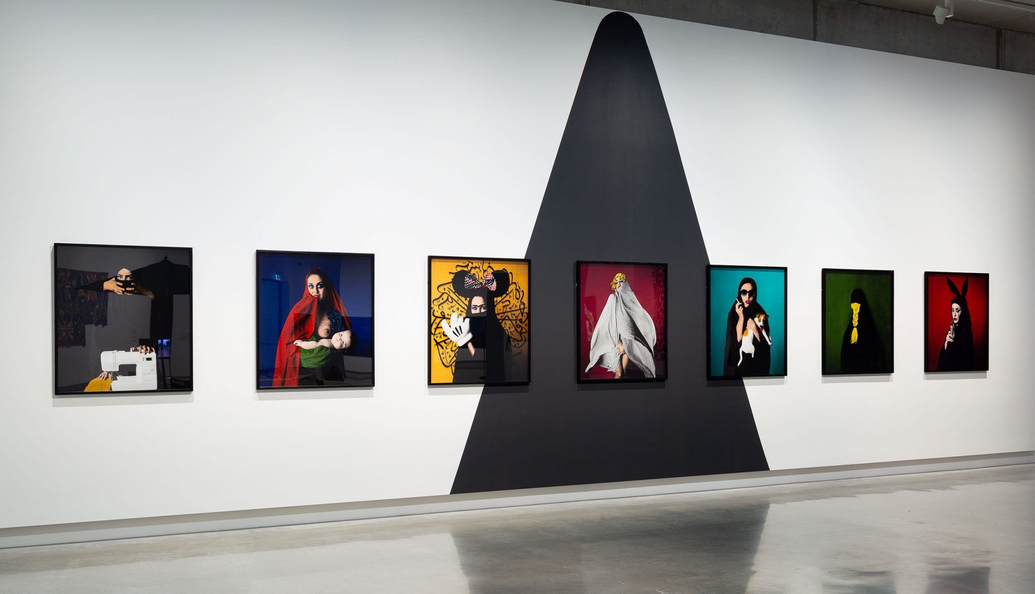 A gallery wall displays seven colorful portrait photographs, each featuring a person in distinct poses and costumes. A large black triangular shape is painted on the white wall behind the artworks. The floor is shiny and reflective.