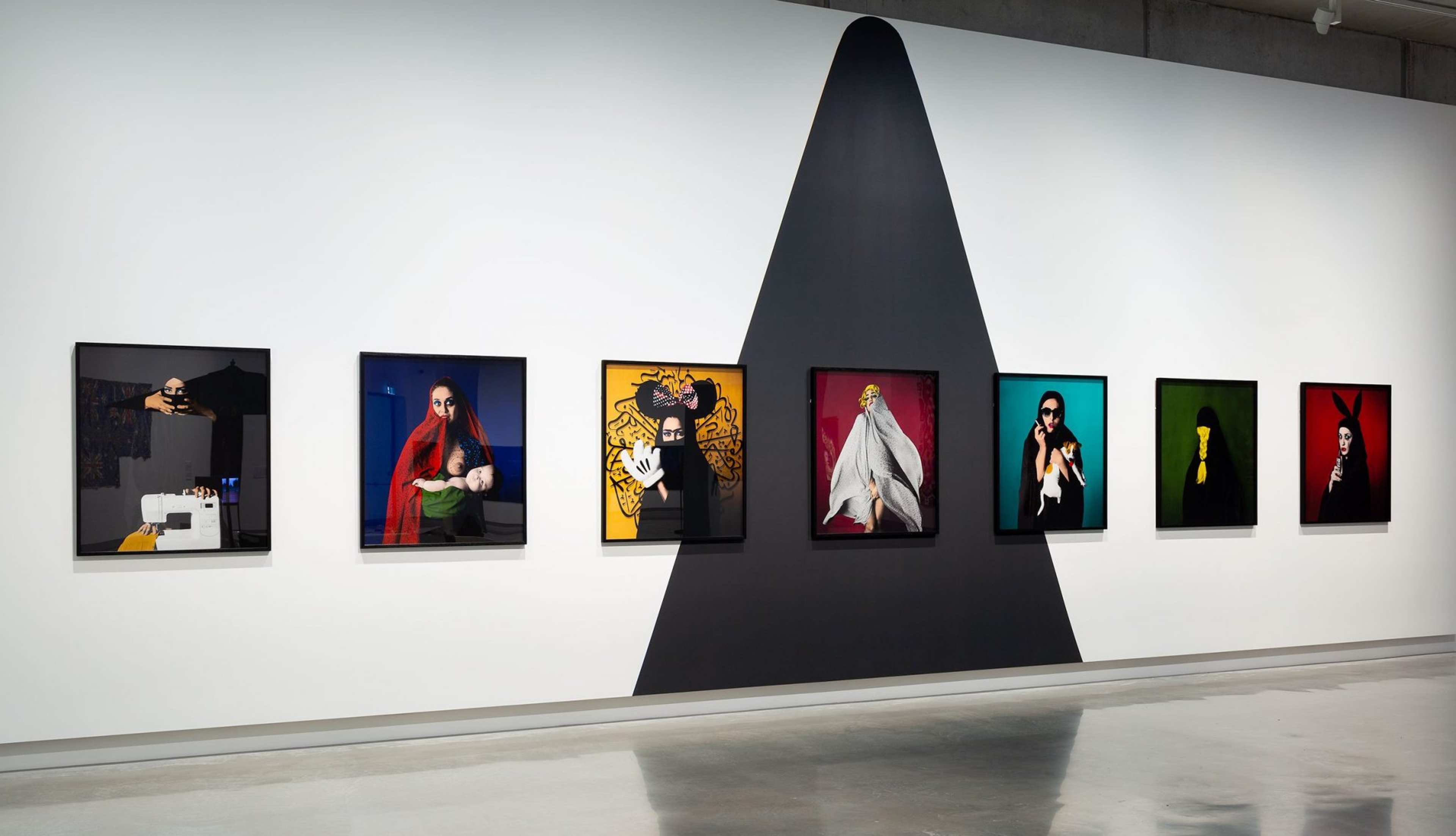 A gallery wall displays seven colorful portrait photographs, each featuring a person in distinct poses and costumes. A large black triangular shape is painted on the white wall behind the artworks. The floor is shiny and reflective.