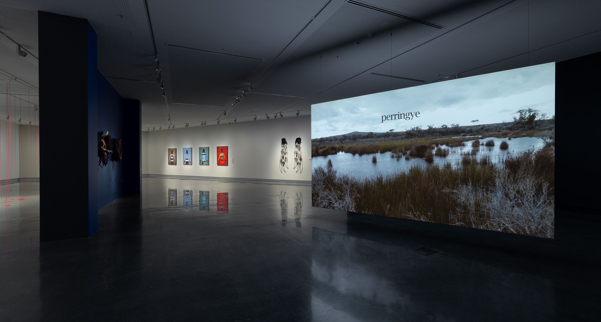 A modern art gallery with polished floors, artworks displayed on white walls, and a large video projection showing a landscape scene with the word perrniŋye on it. The space is dimly lit, creating a calm atmosphere.