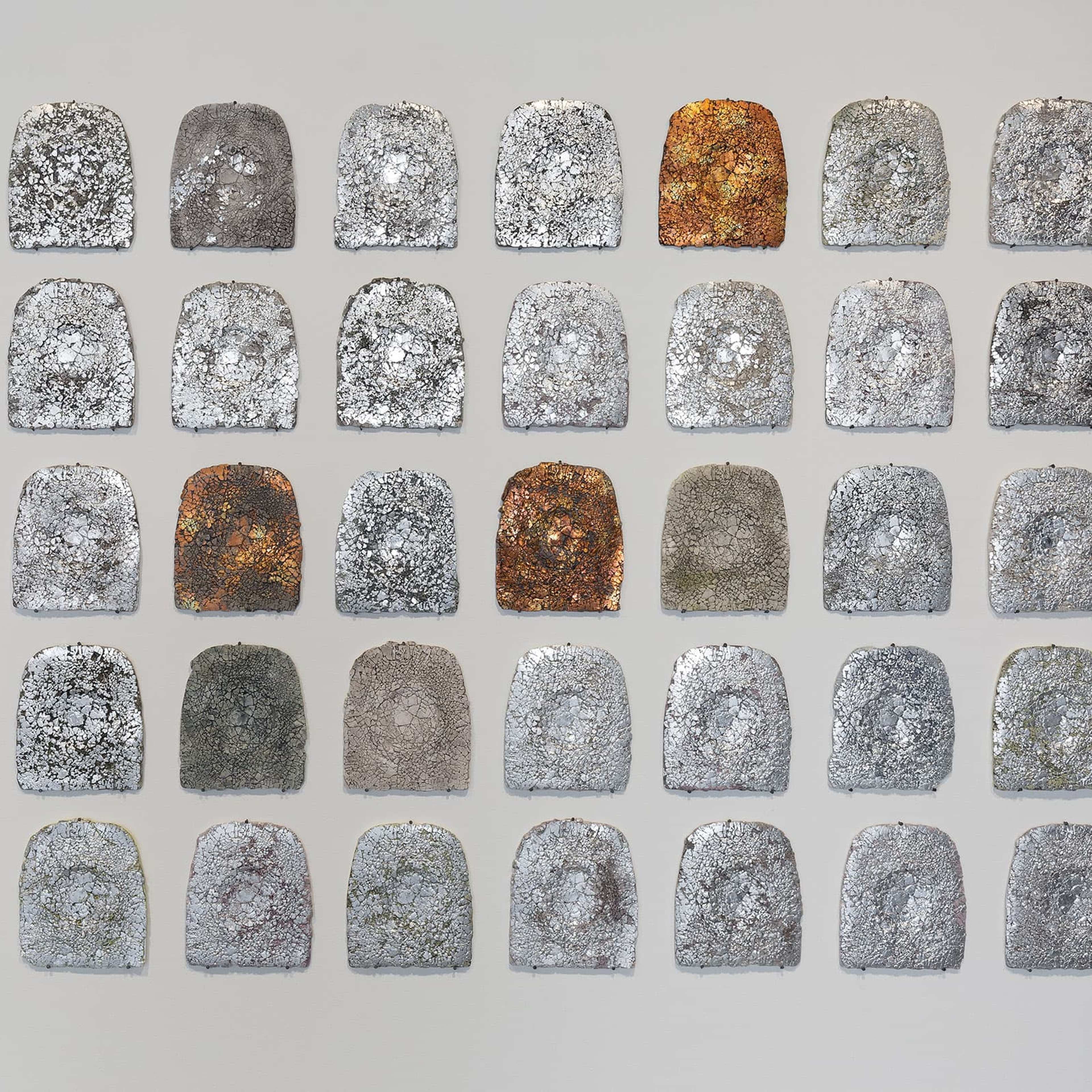 A grid of 40 tombstone-shaped plaques, each with a unique texture and color ranging from gray and silver to rust and brown, arranged in five rows on a light gray background.