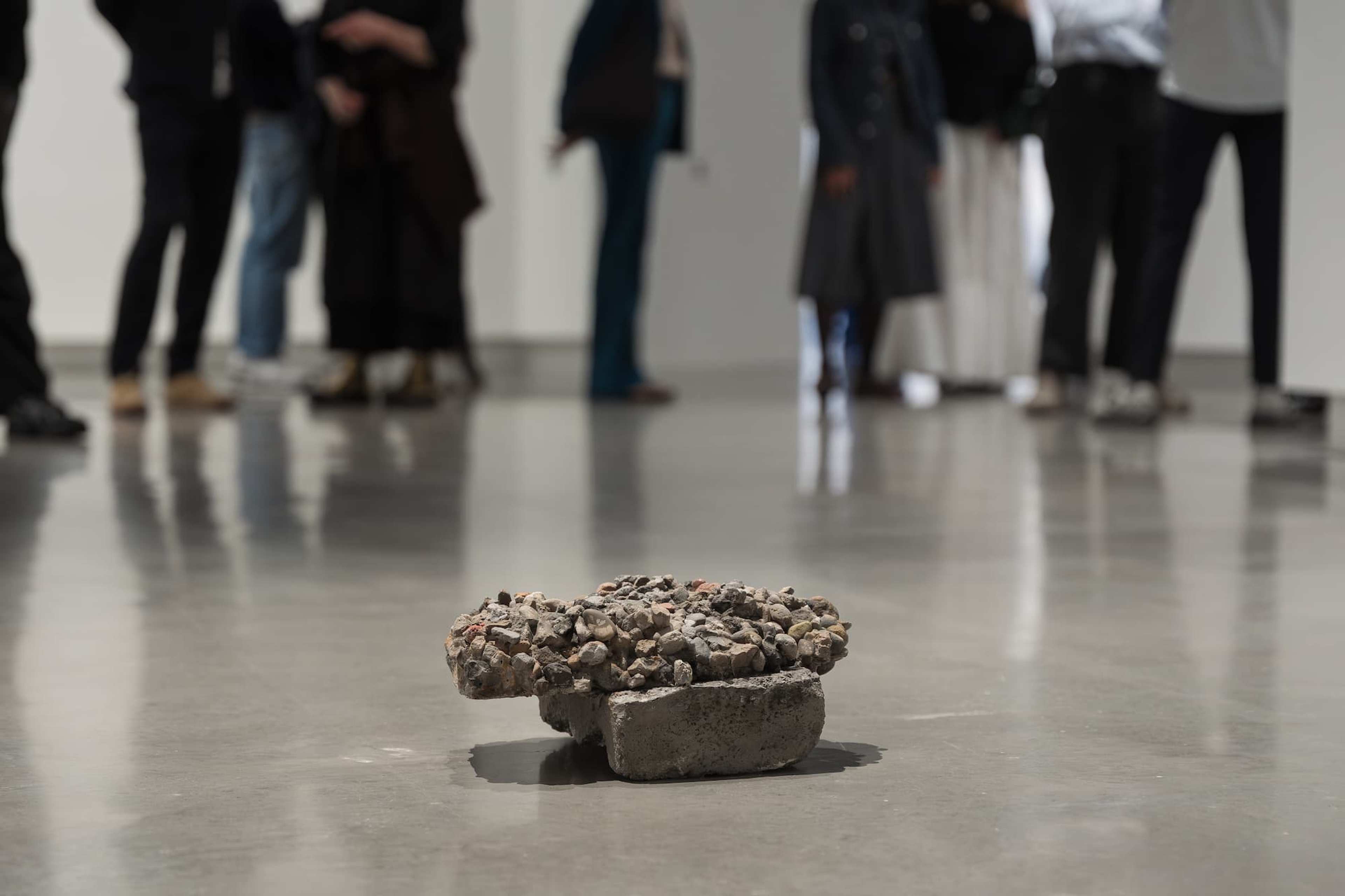 A small, rough piece of concrete with gravel sits on a shiny gallery floor, while a group of people in blurred focus stand and talk in the background.