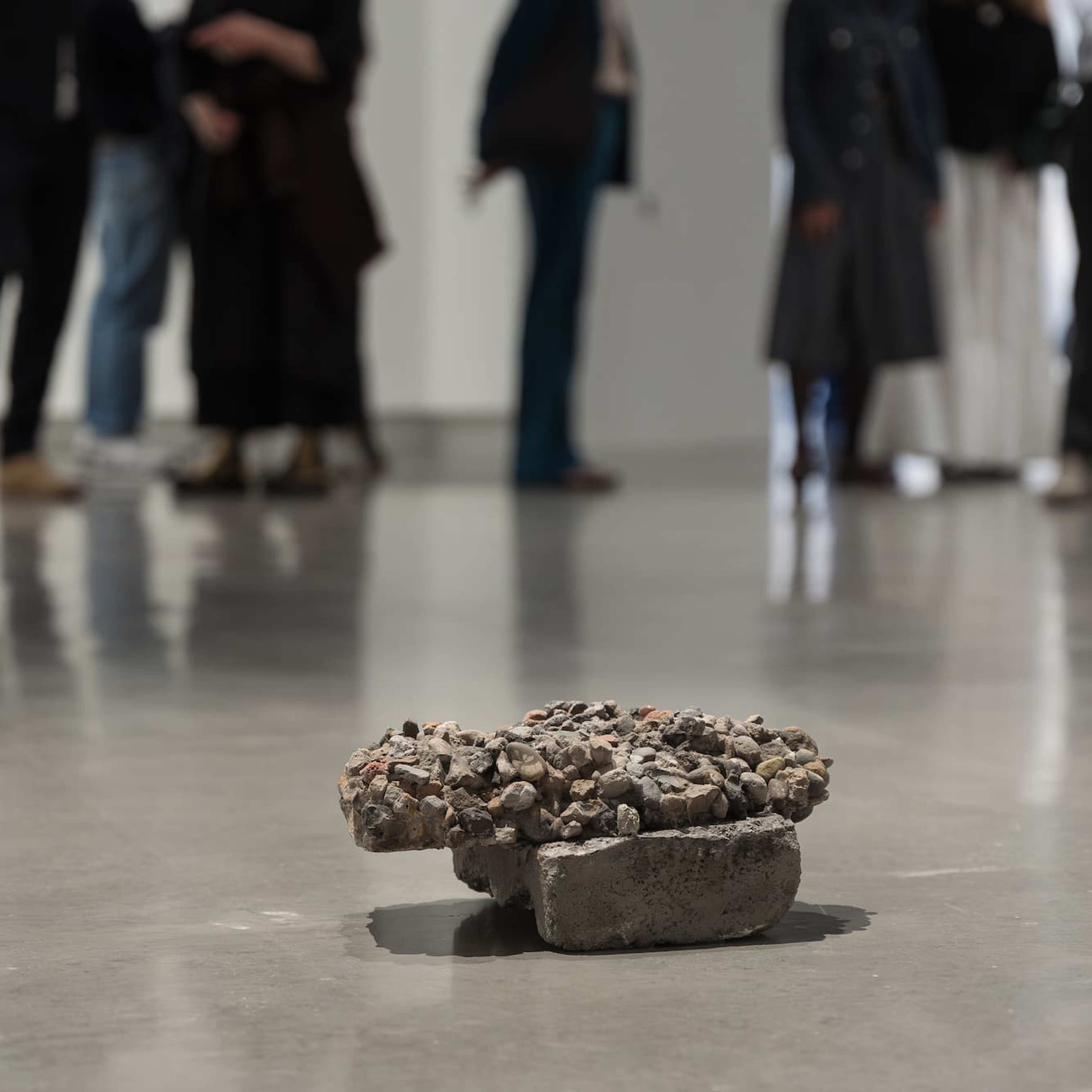 A small, rough piece of concrete with gravel sits on a shiny gallery floor, while a group of people in blurred focus stand and talk in the background.