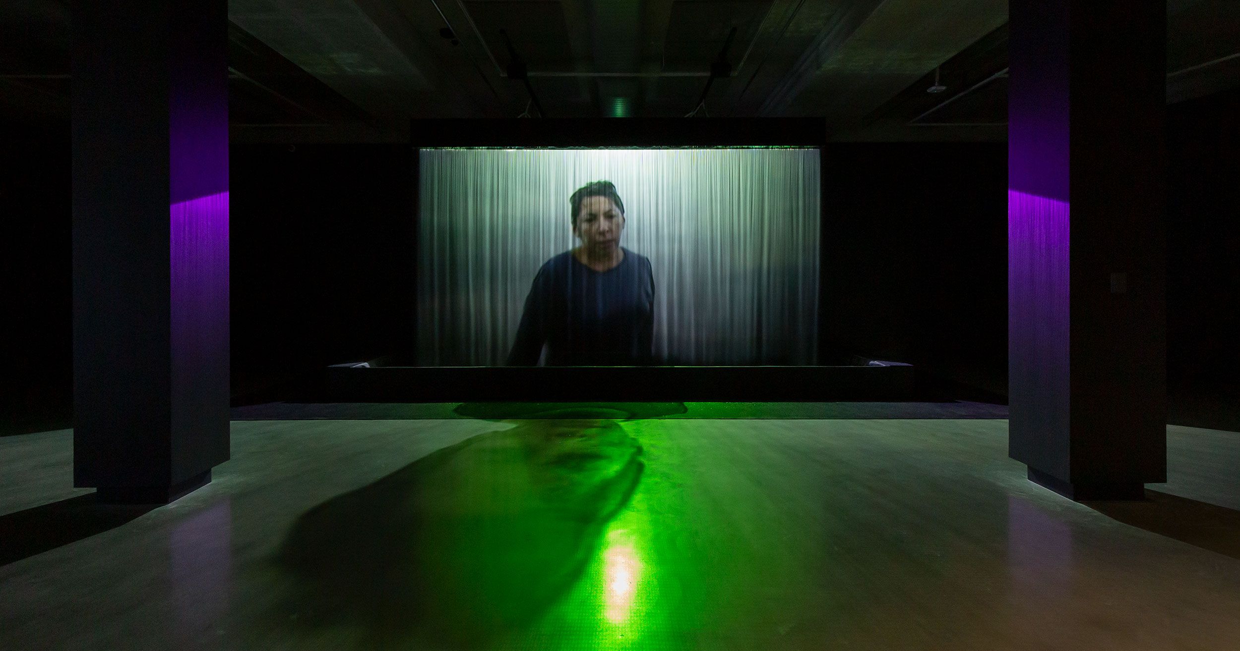 A dimly lit room features a large curtained screen showing a person standing, with green and purple lights casting shadows and reflections on the floor. The atmosphere is mysterious and immersive.