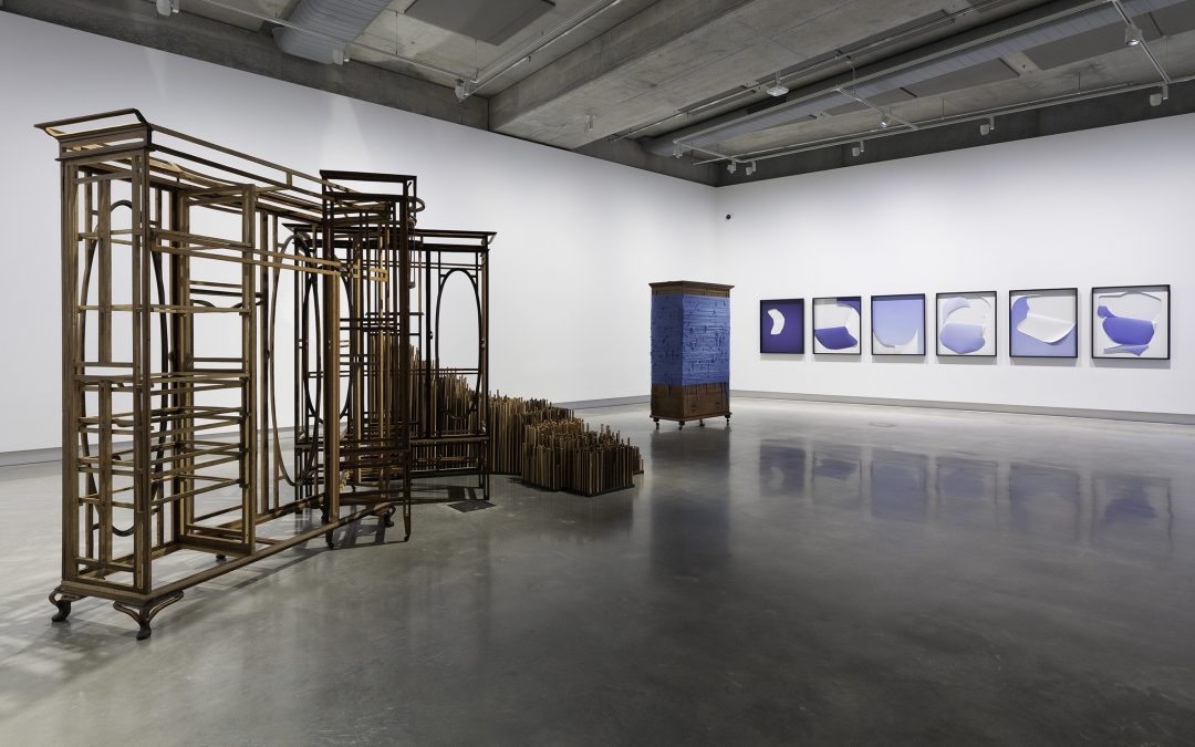A modern art gallery with sculptural metal and wood structures on the left and center, and a series of six framed abstract blue and white prints displayed on the far white wall. The floor is polished concrete.