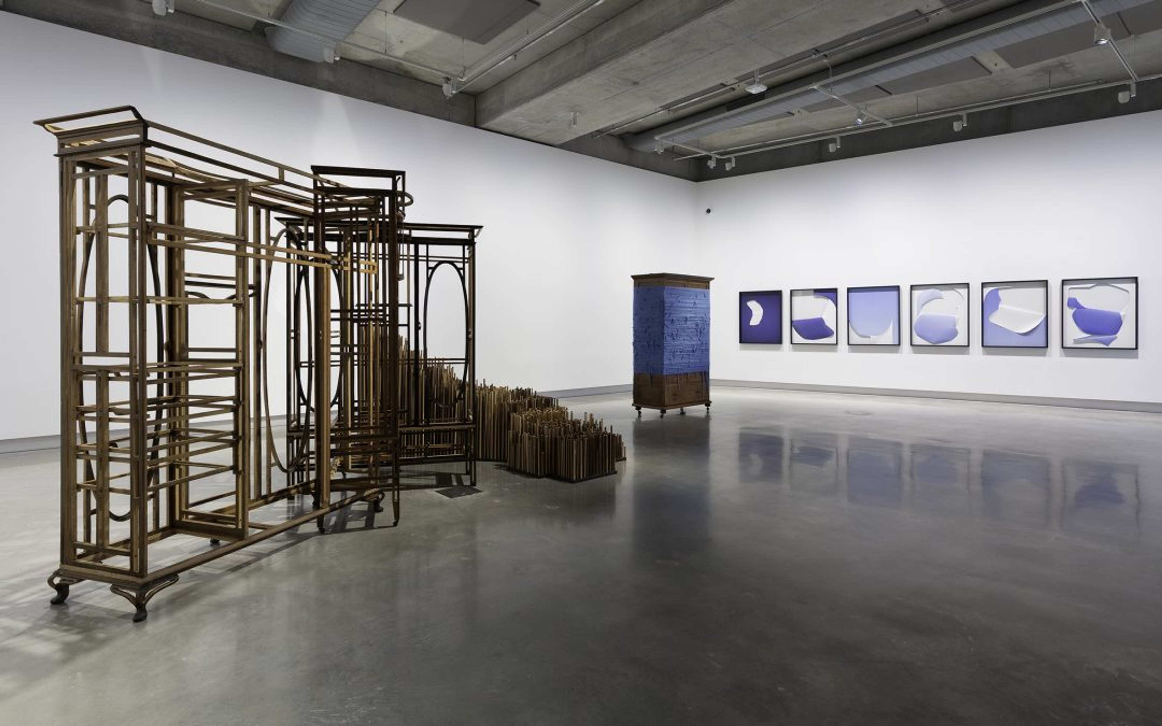 A modern art gallery with sculptural metal and wood structures on the left and center, and a series of six framed abstract blue and white prints displayed on the far white wall. The floor is polished concrete.