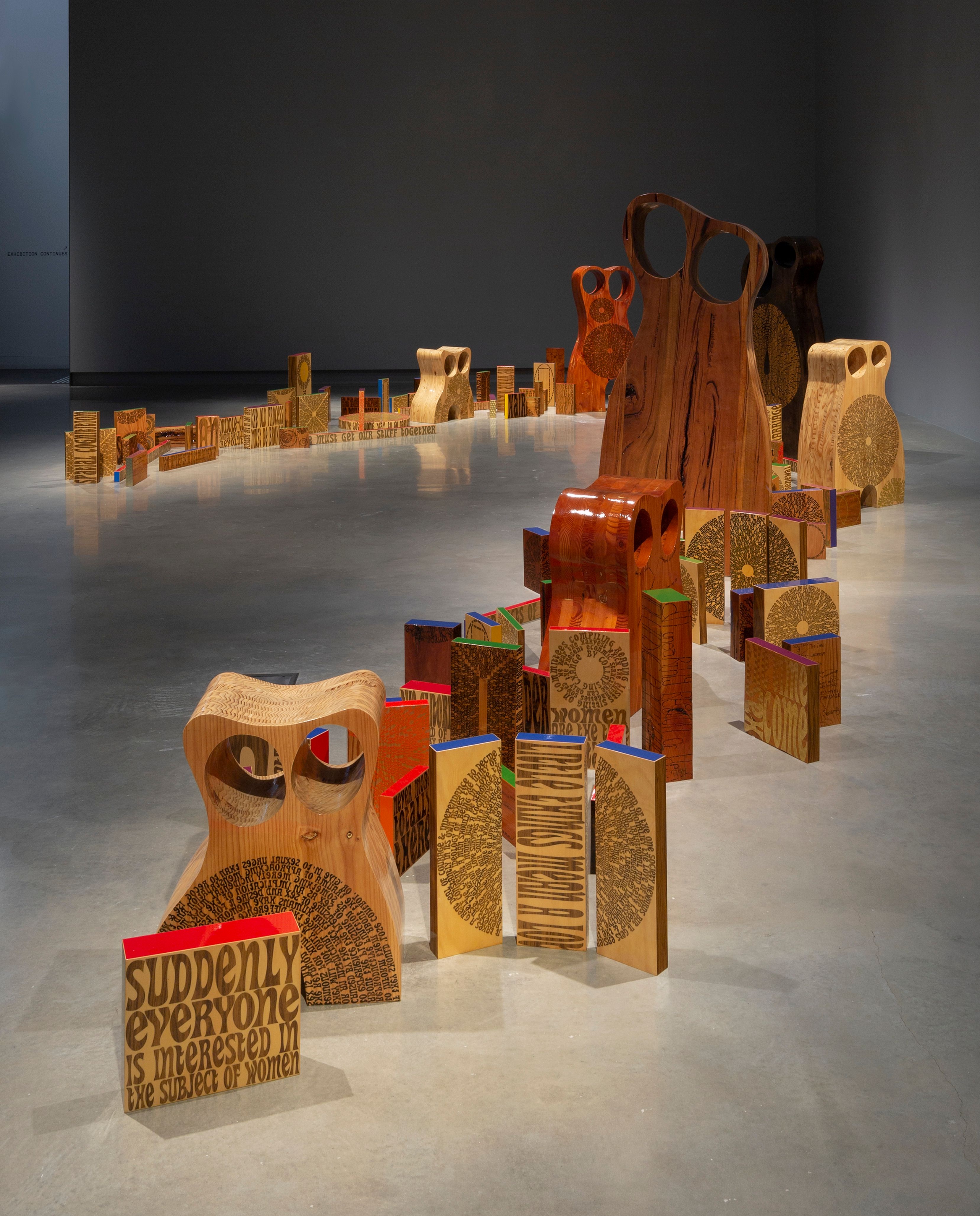 Irregularly shaped wooden sculptures with text engraved, lined up across a gallery floor.