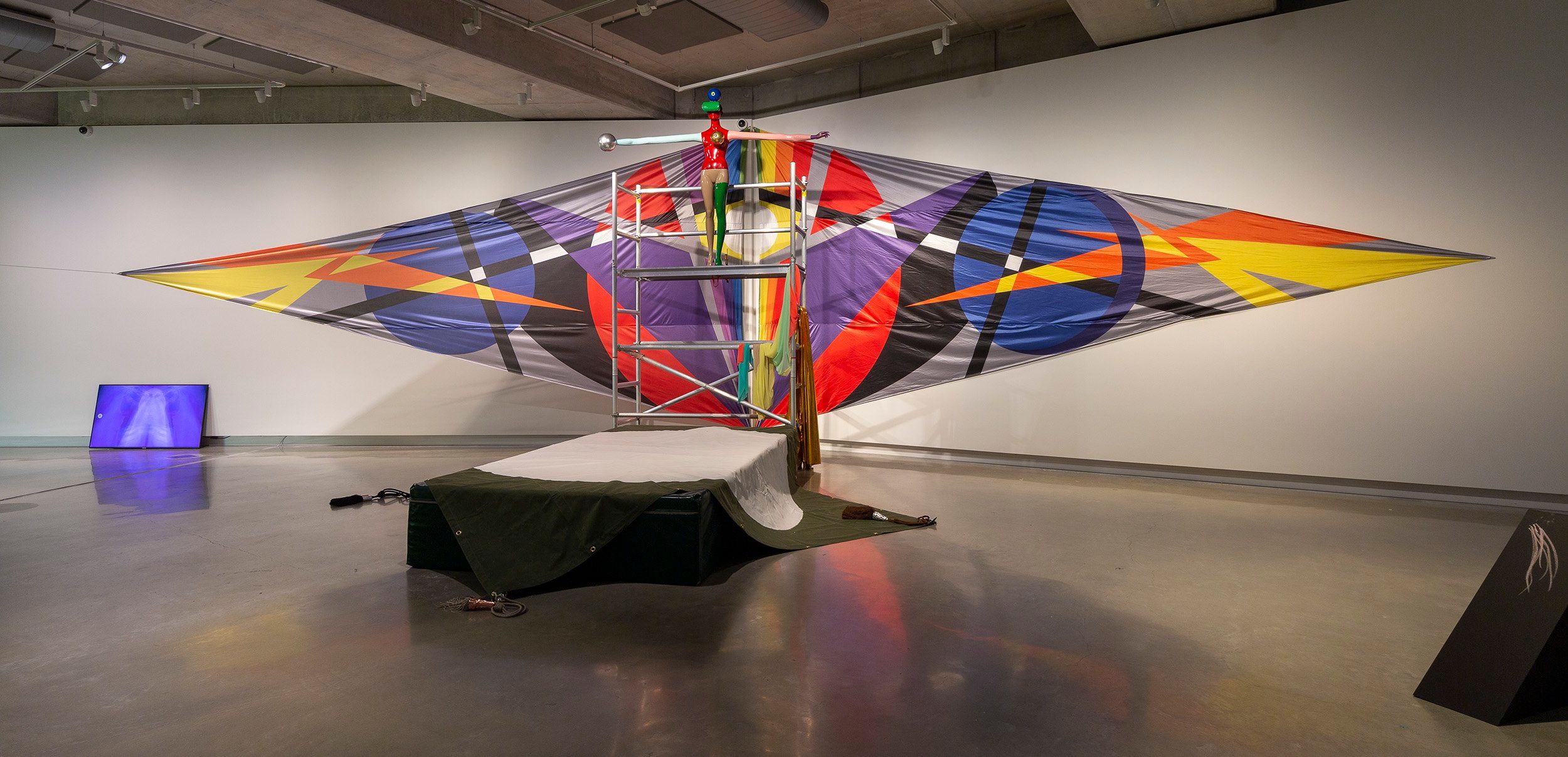 Large colorful geometric sculpture resembling an abstract kite, with a central frame and beams supporting fabric in red, purple, orange, yellow, and black, displayed in a modern gallery space with neutral walls and polished floors.