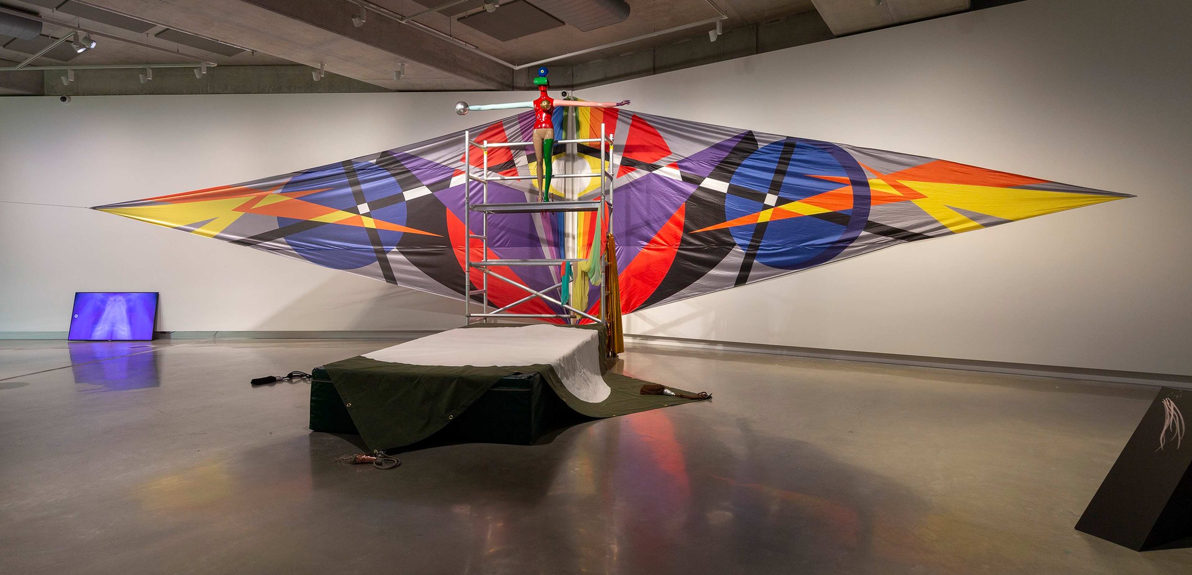 Large colorful geometric sculpture resembling an abstract kite, with a central frame and beams supporting fabric in red, purple, orange, yellow, and black, displayed in a modern gallery space with neutral walls and polished floors.
