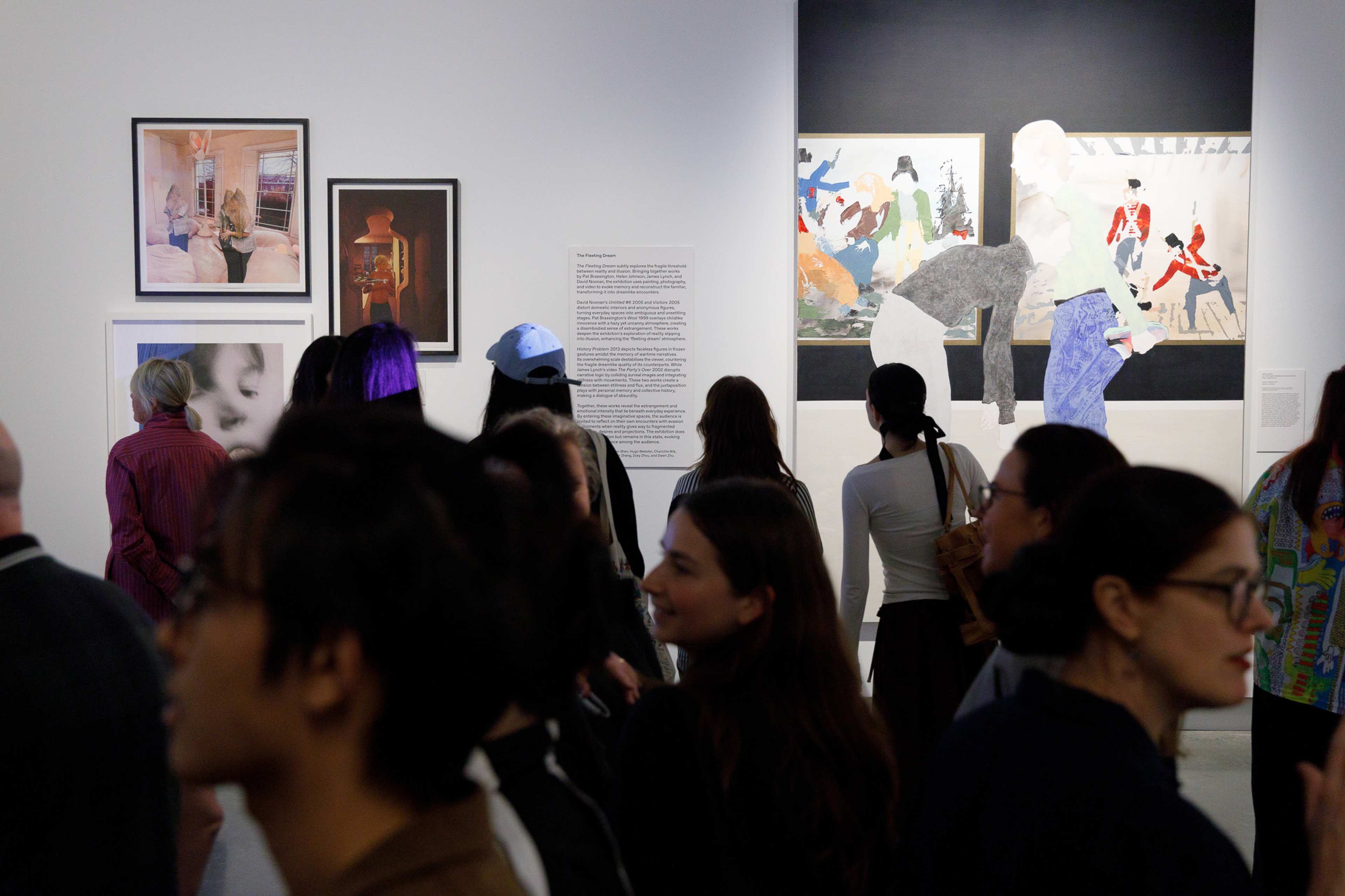 A crowd of people view various colorful paintings and artwork displayed on the walls of a brightly lit art gallery. Some visitors are closely examining the art while others are engaged in conversation.