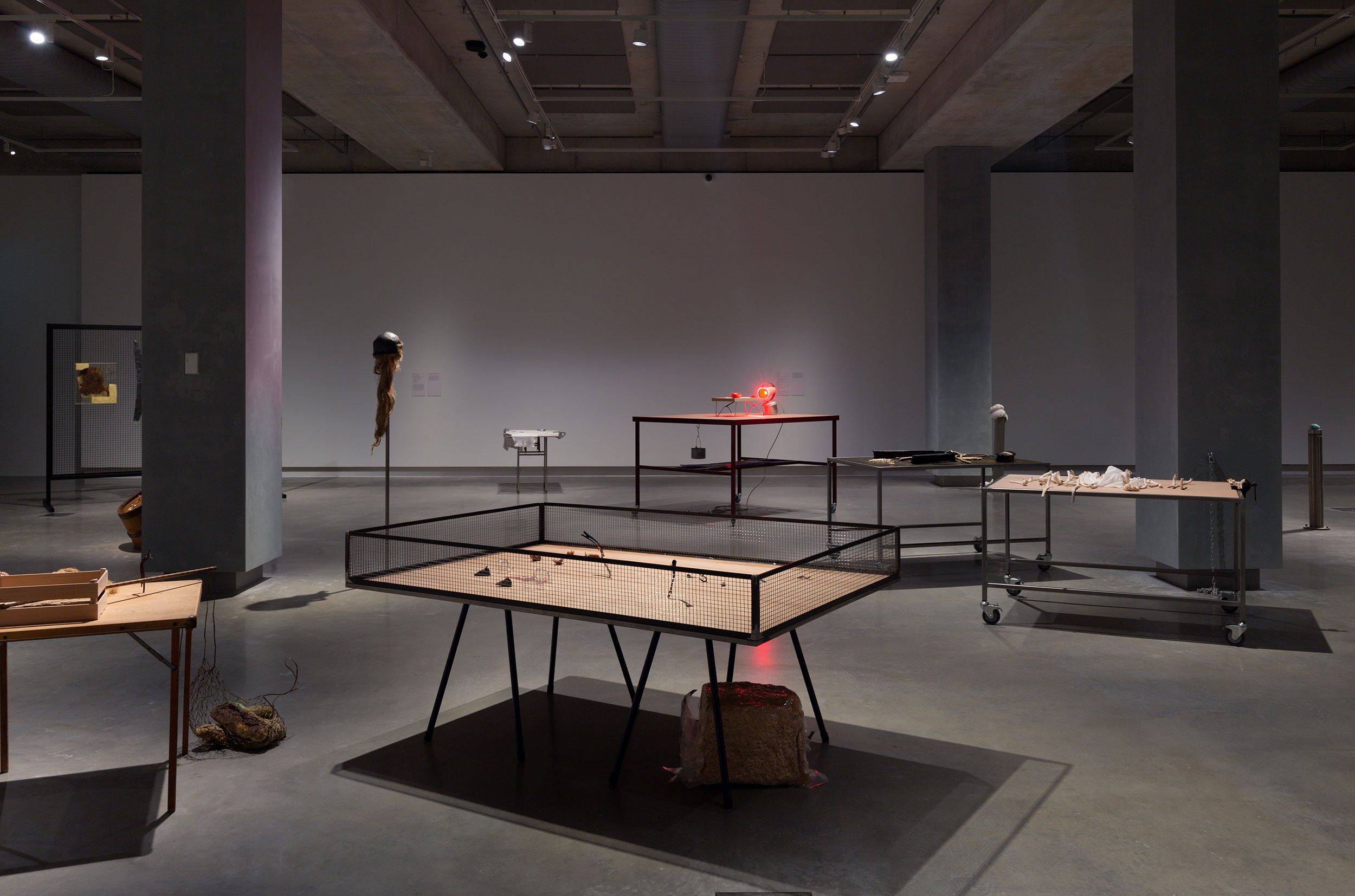 A contemporary art gallery with sculptures and mixed media installations displayed on tables and stands, under ceiling spotlights in a spacious, industrial-style room with concrete floors and pillars.
