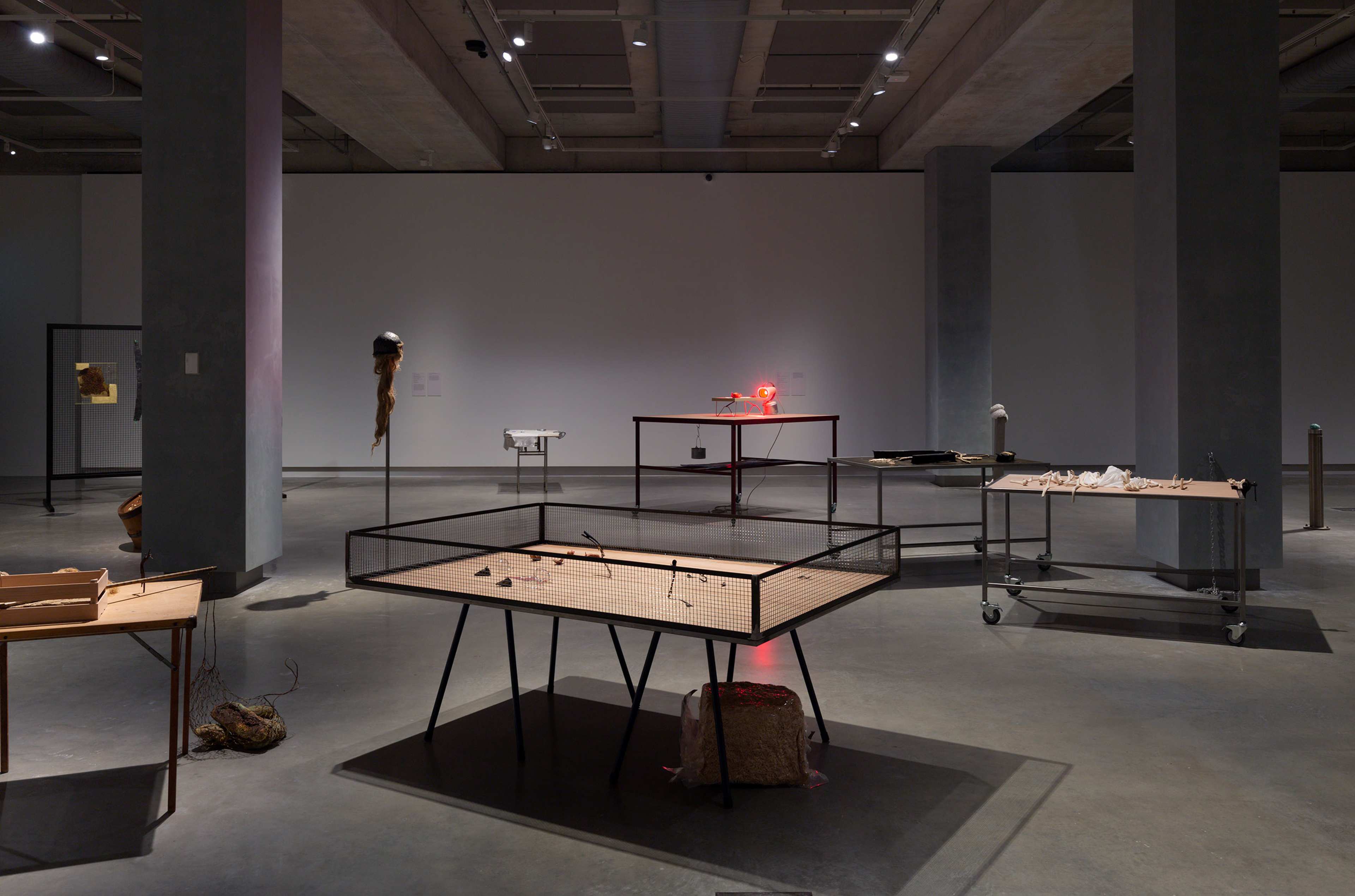 A contemporary art gallery with sculptures and mixed media installations displayed on tables and stands, under ceiling spotlights in a spacious, industrial-style room with concrete floors and pillars.