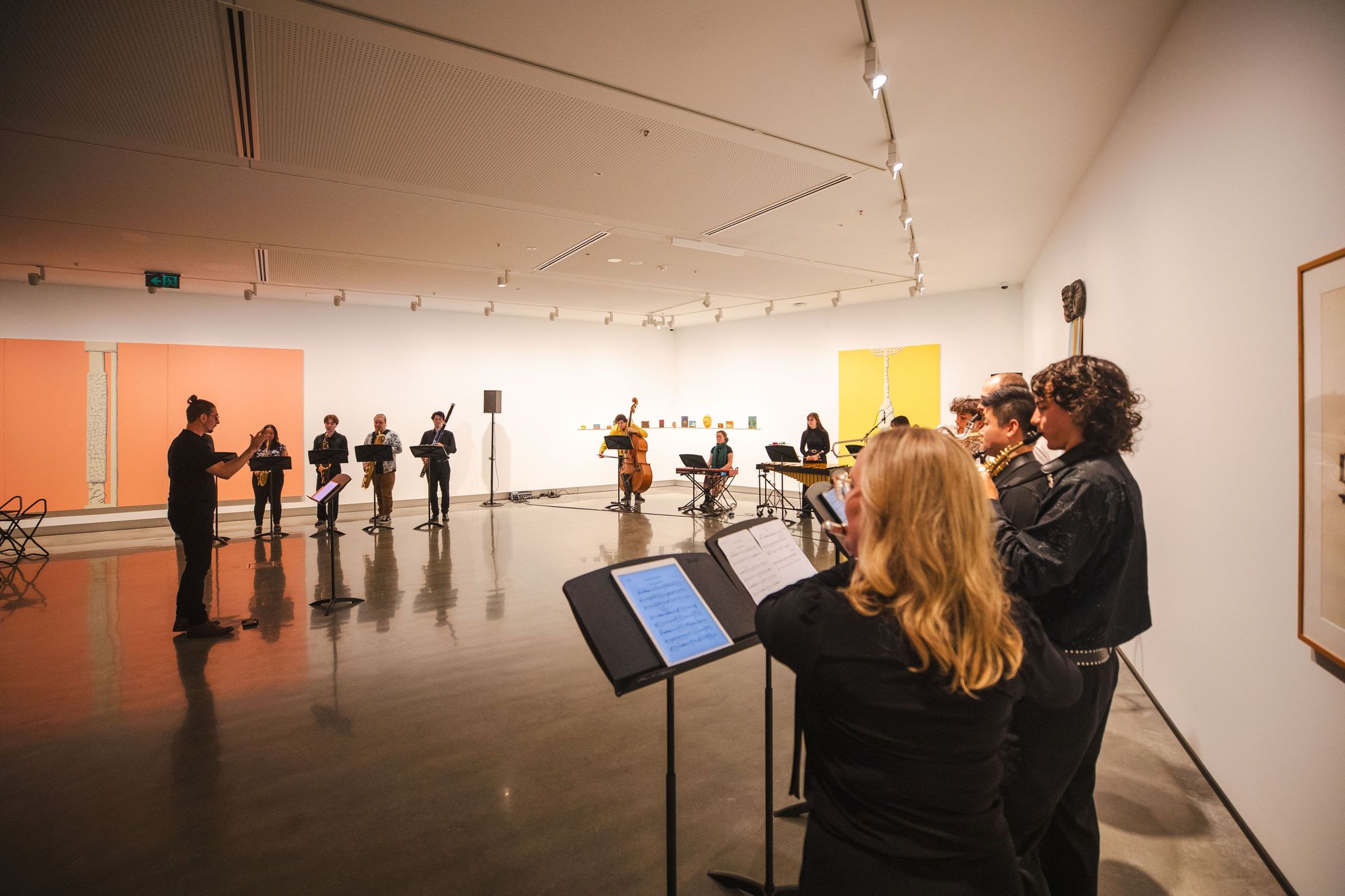 A group of musicians perform indoors in an art gallery, standing in a semicircle with music stands. Artworks are displayed on the walls, and the room is brightly lit with spotlights on the ceiling.