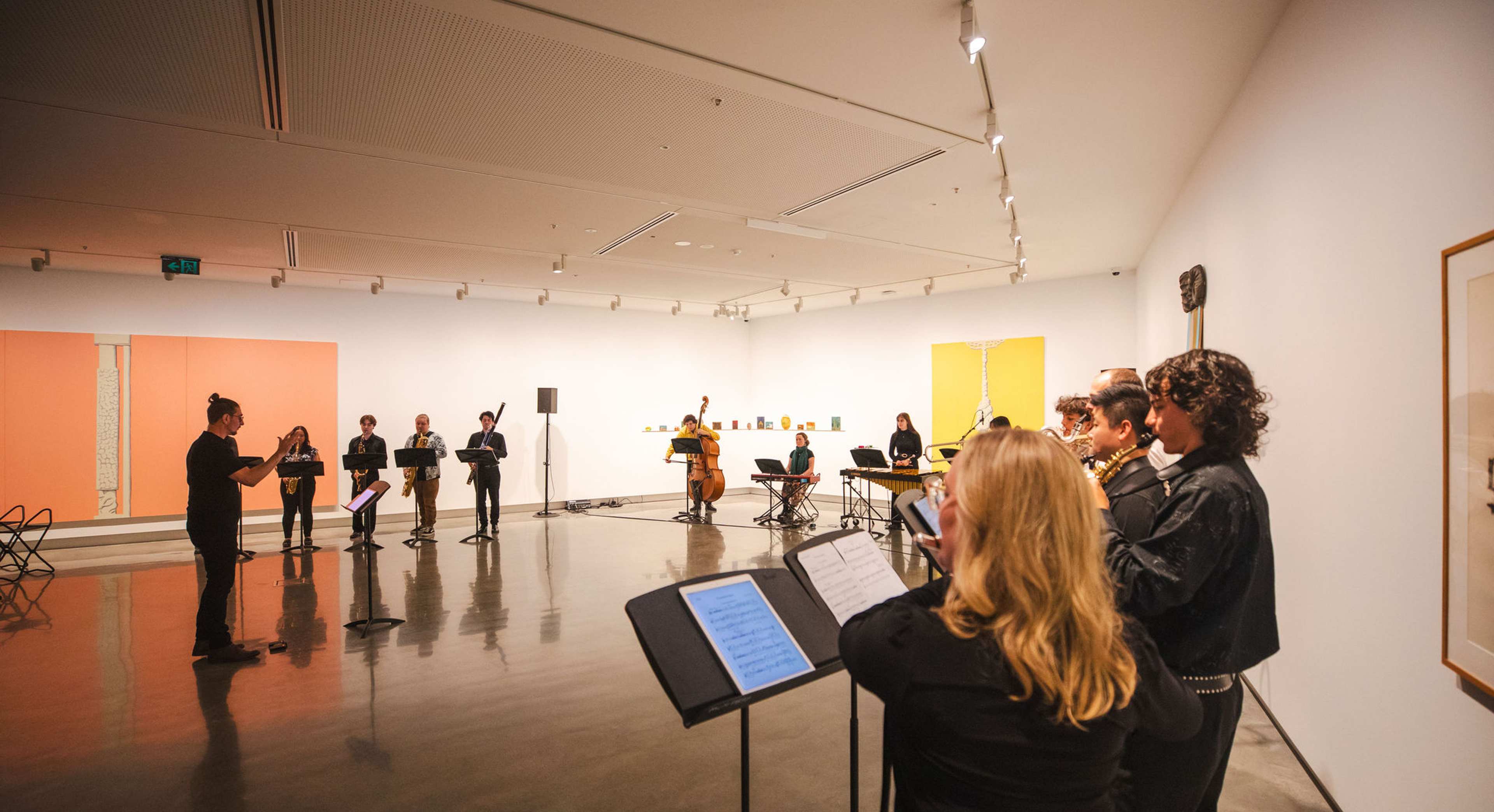 A group of musicians perform indoors in an art gallery, standing in a semicircle with music stands. Artworks are displayed on the walls, and the room is brightly lit with spotlights on the ceiling.