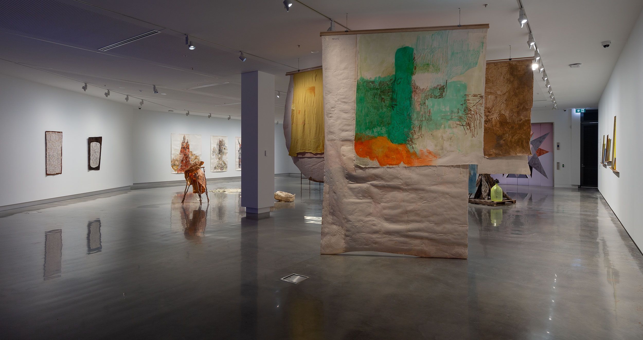 A modern art gallery with abstract paintings and mixed-media fabric pieces hanging from the ceiling, displayed in a spacious room with polished floors and white walls. The space is well-lit with track lighting.