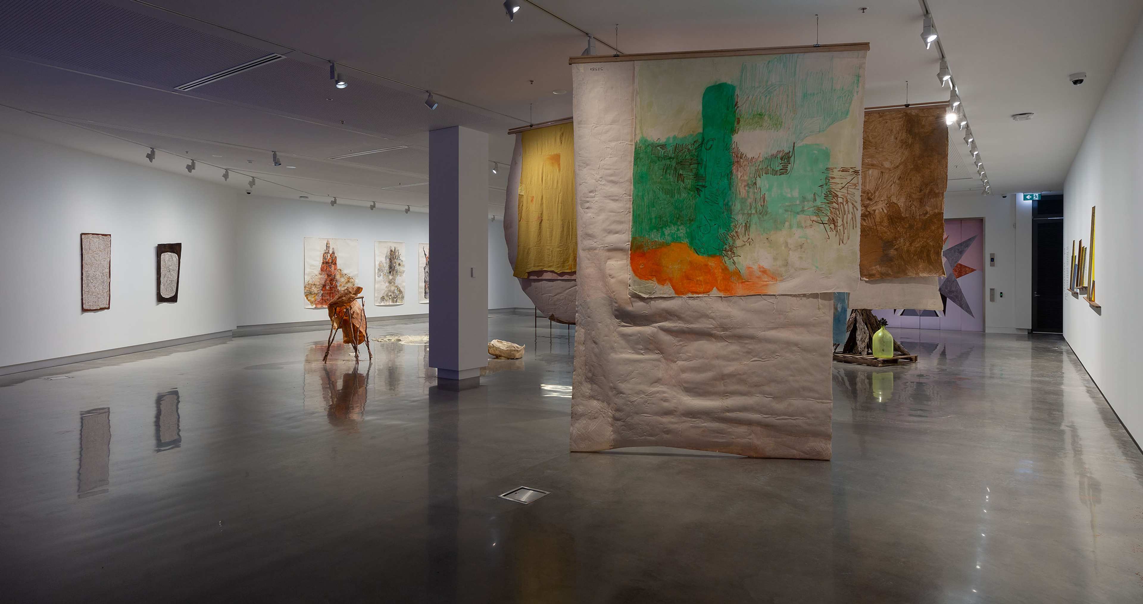 A modern art gallery with abstract paintings and mixed-media fabric pieces hanging from the ceiling, displayed in a spacious room with polished floors and white walls. The space is well-lit with track lighting.