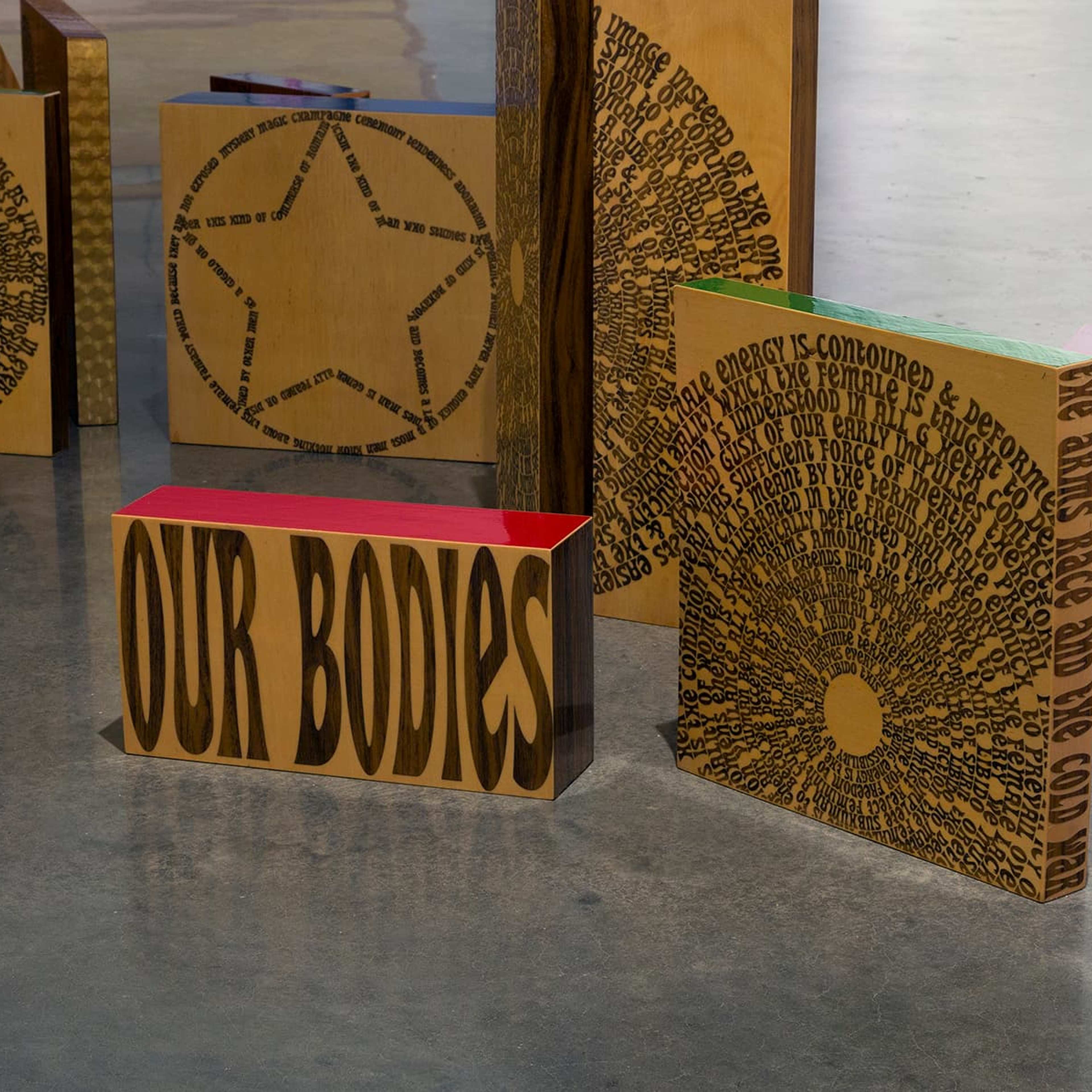 Wooden panels with engraved text and designs, including star shapes and circular patterns, are arranged upright on a concrete floor. One panel in the foreground prominently displays the words “OUR BODIES” in large letters.