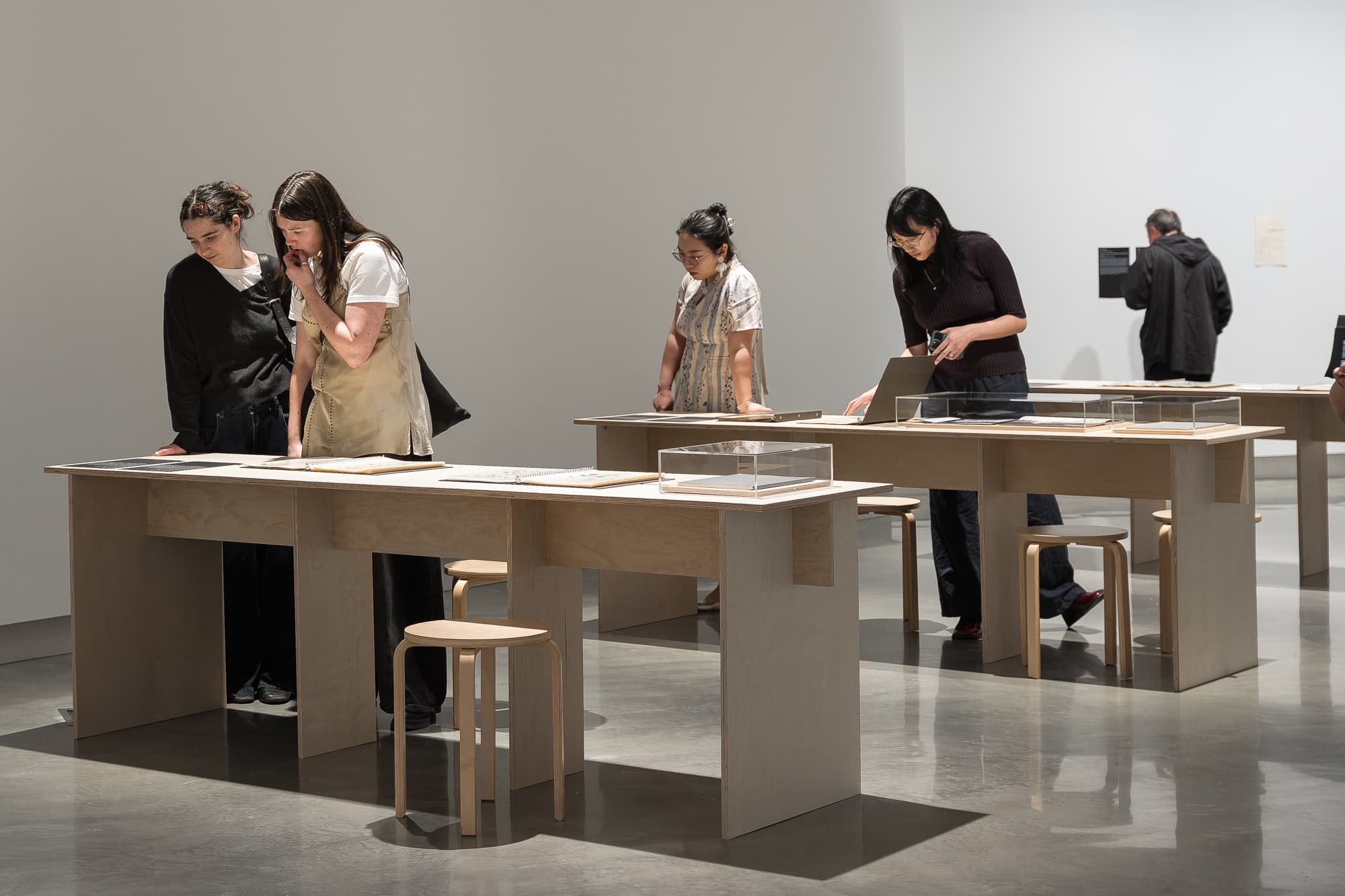 Five people examine documents and displays on light wooden tables in a modern, minimalist gallery with white walls and polished floors. Some are closely reading, while others interact quietly with the exhibits.