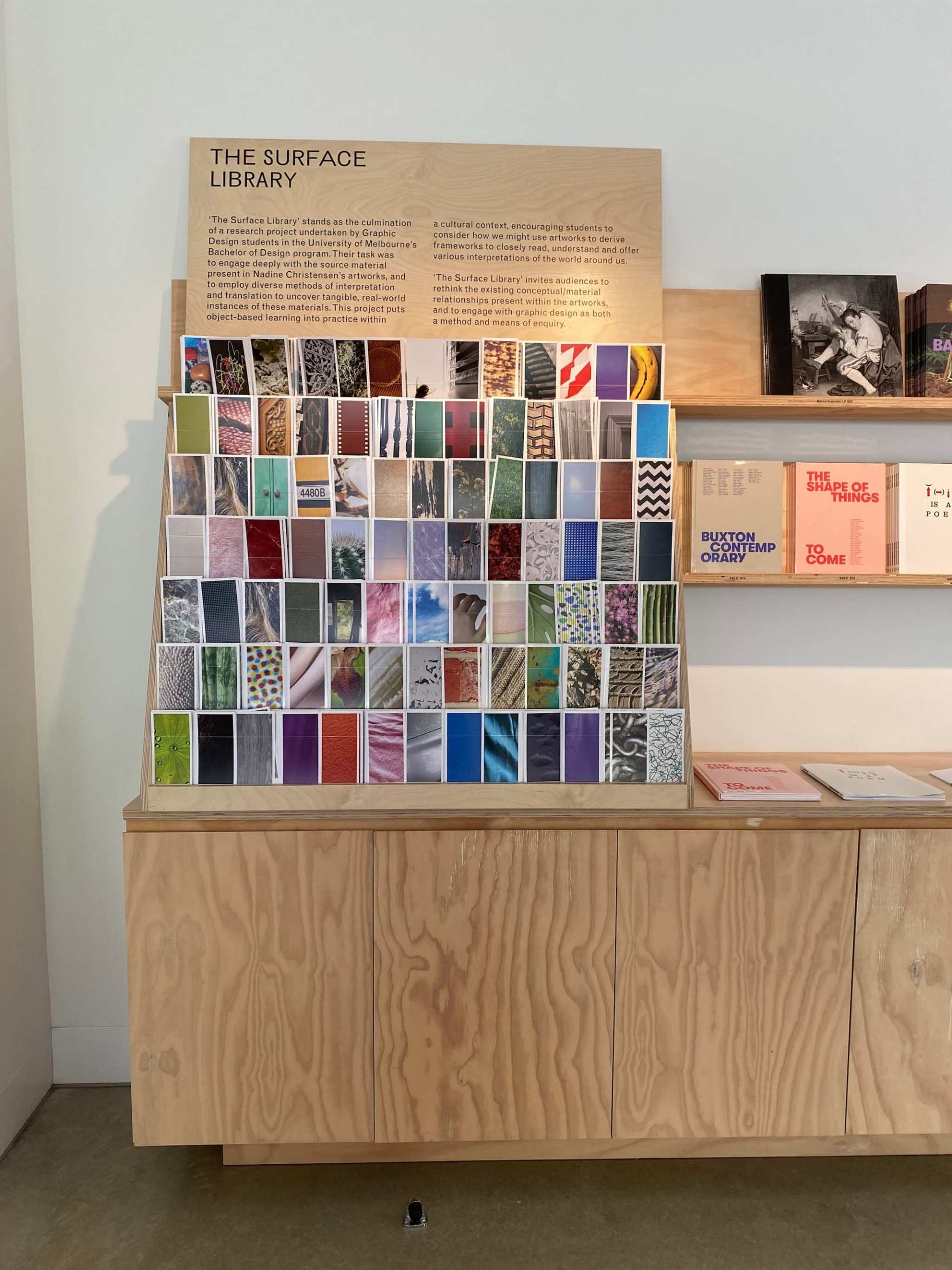 A wooden display labeled The Surface Library shows a grid of colorful material samples, each with a unique texture or pattern, in a modern, minimalistic room with books on shelves beside and below the display.