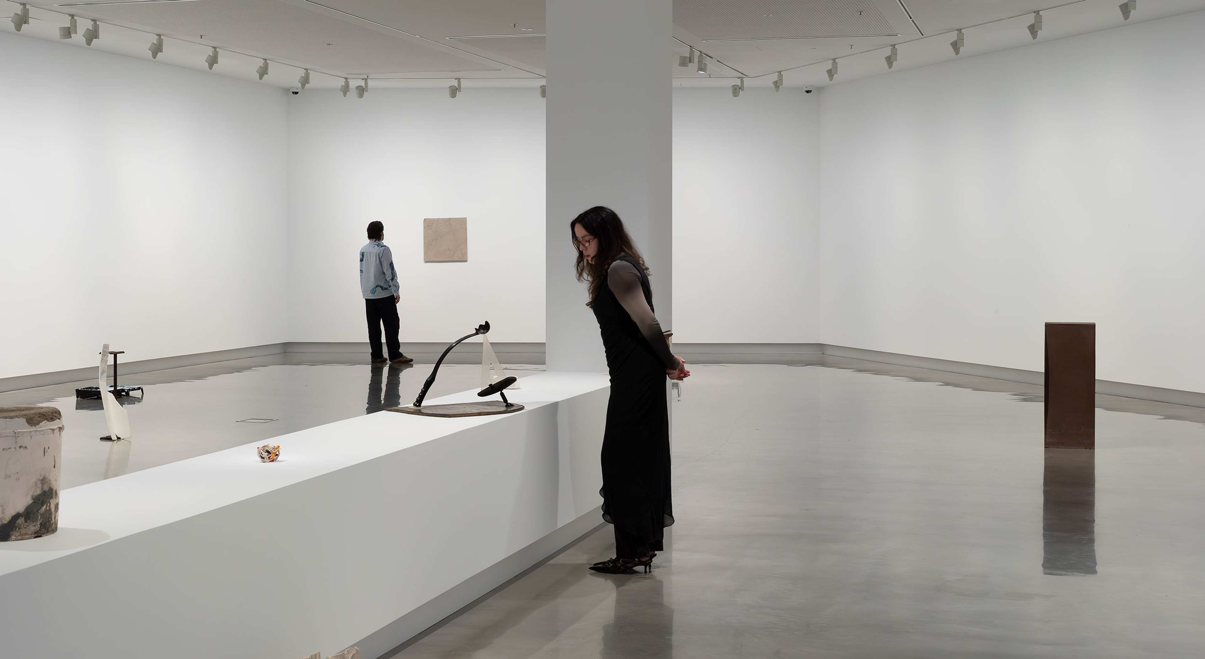 A person examines minimalist sculptures on a white display in a modern, spacious art gallery with polished floors. In the background, another person views artwork on the far wall. The space is brightly lit and minimalistic.