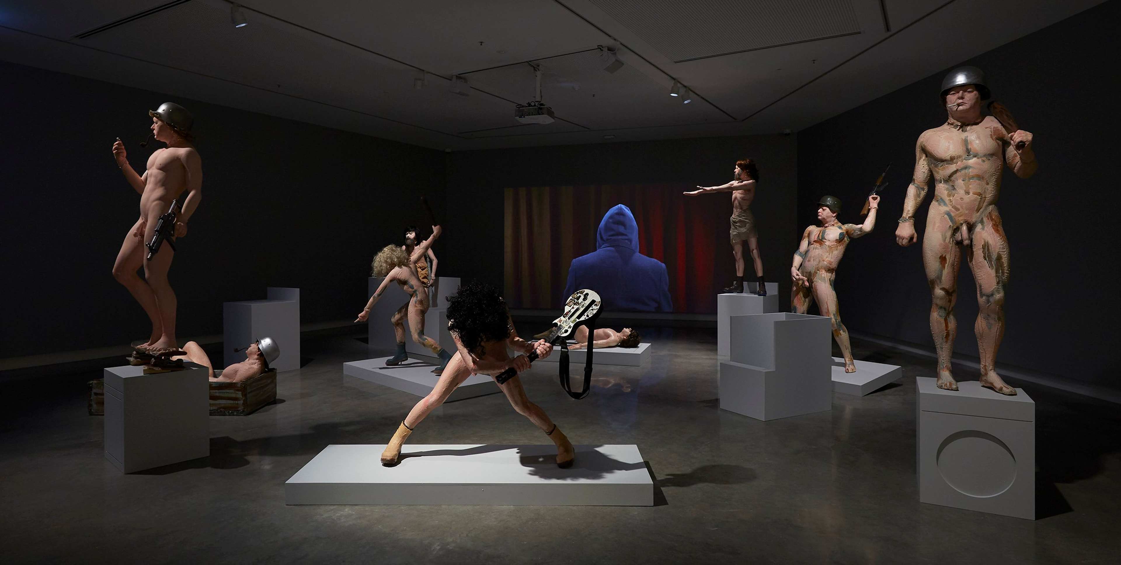 A dimly lit gallery displays several life-sized nude sculptures, some wearing helmets and holding weapons. Figures stand or pose on various pedestals, with a video projection of a hooded person on the far wall.