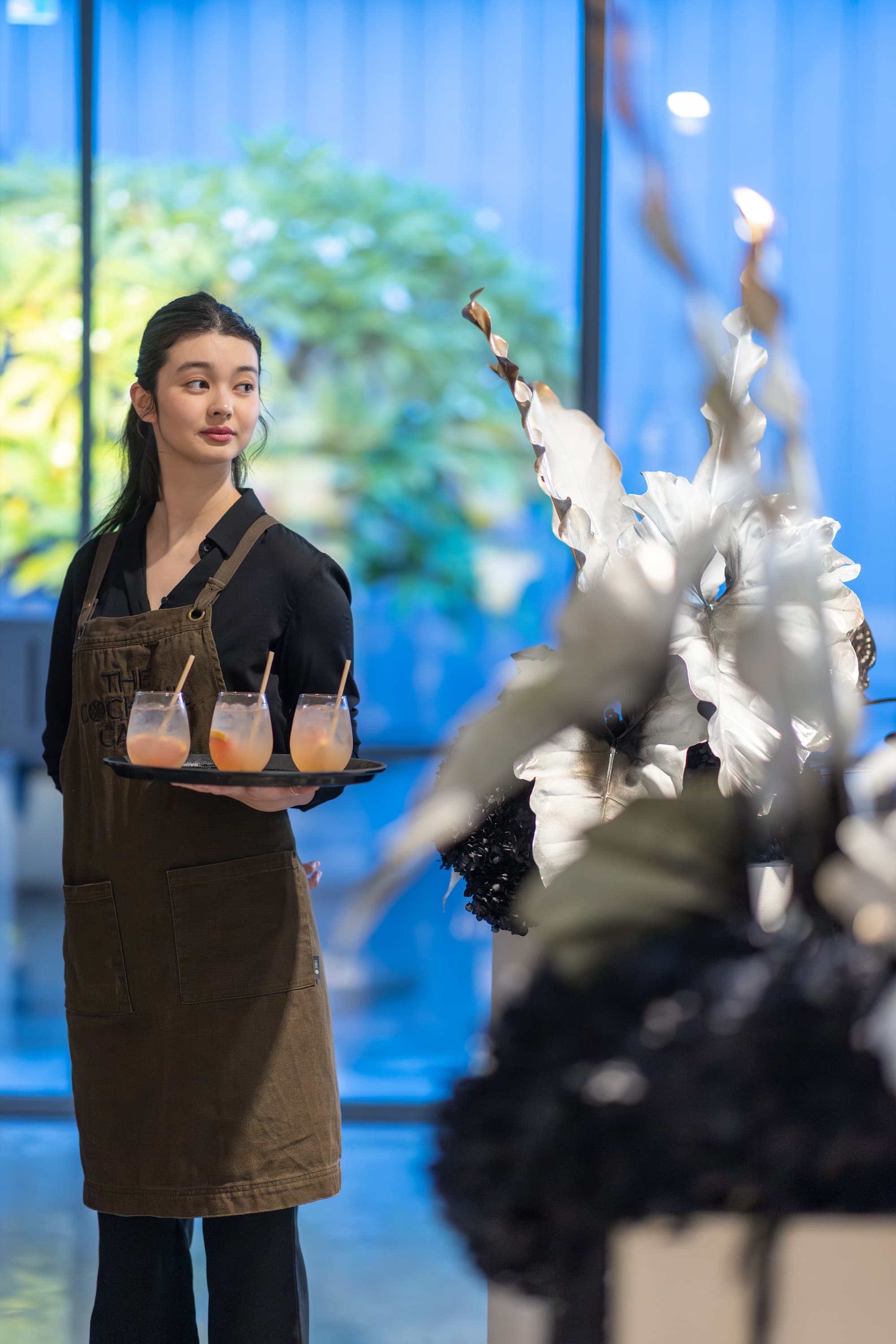 A person in a dark apron stands by a window holding a tray with three drinks