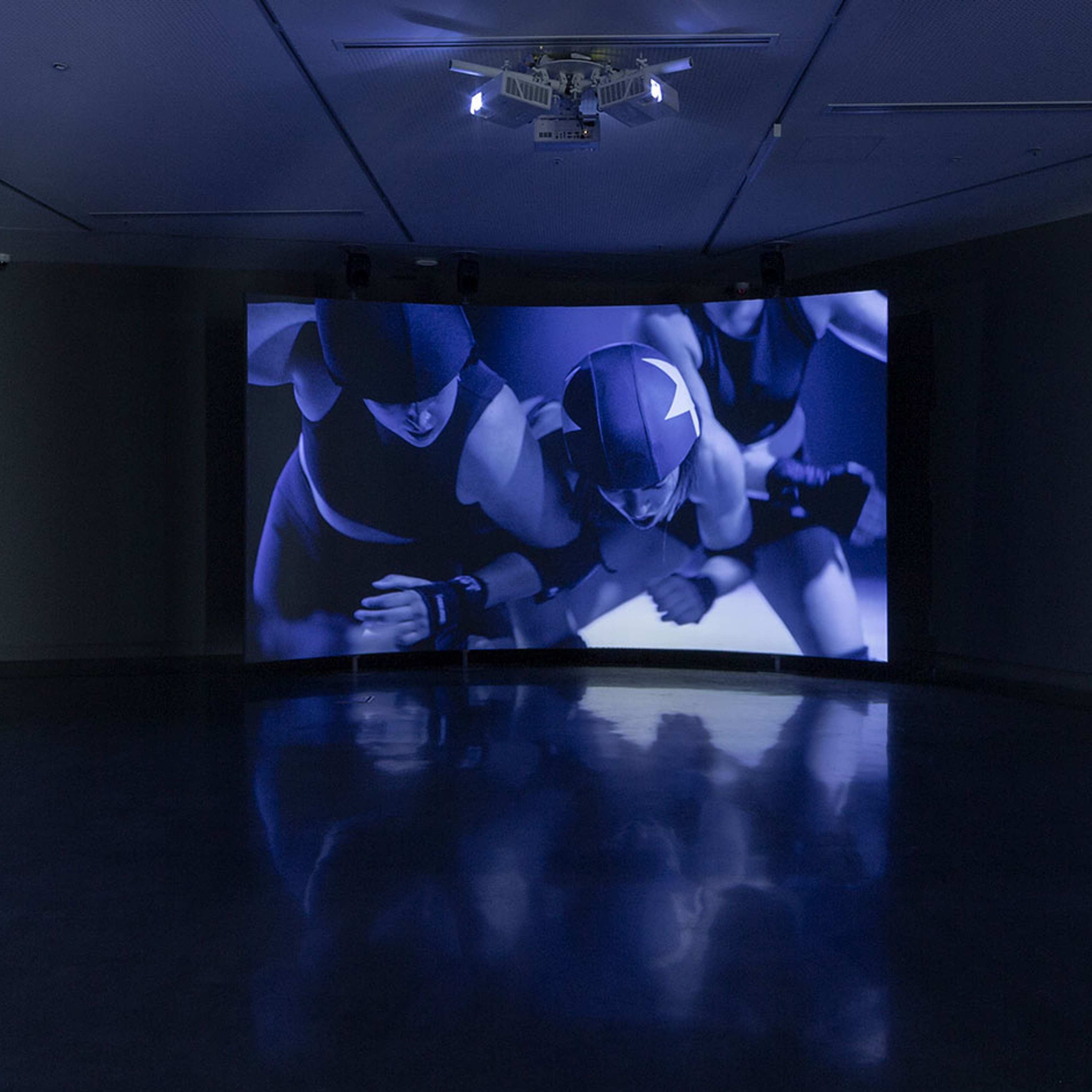 A dark gallery features a large screen displaying three athletes in helmets and uniforms, appearing to wrestle or play a contact sport. Panels curve towards the screen, and the room is dimly lit with blue tones reflected on the shiny floor.