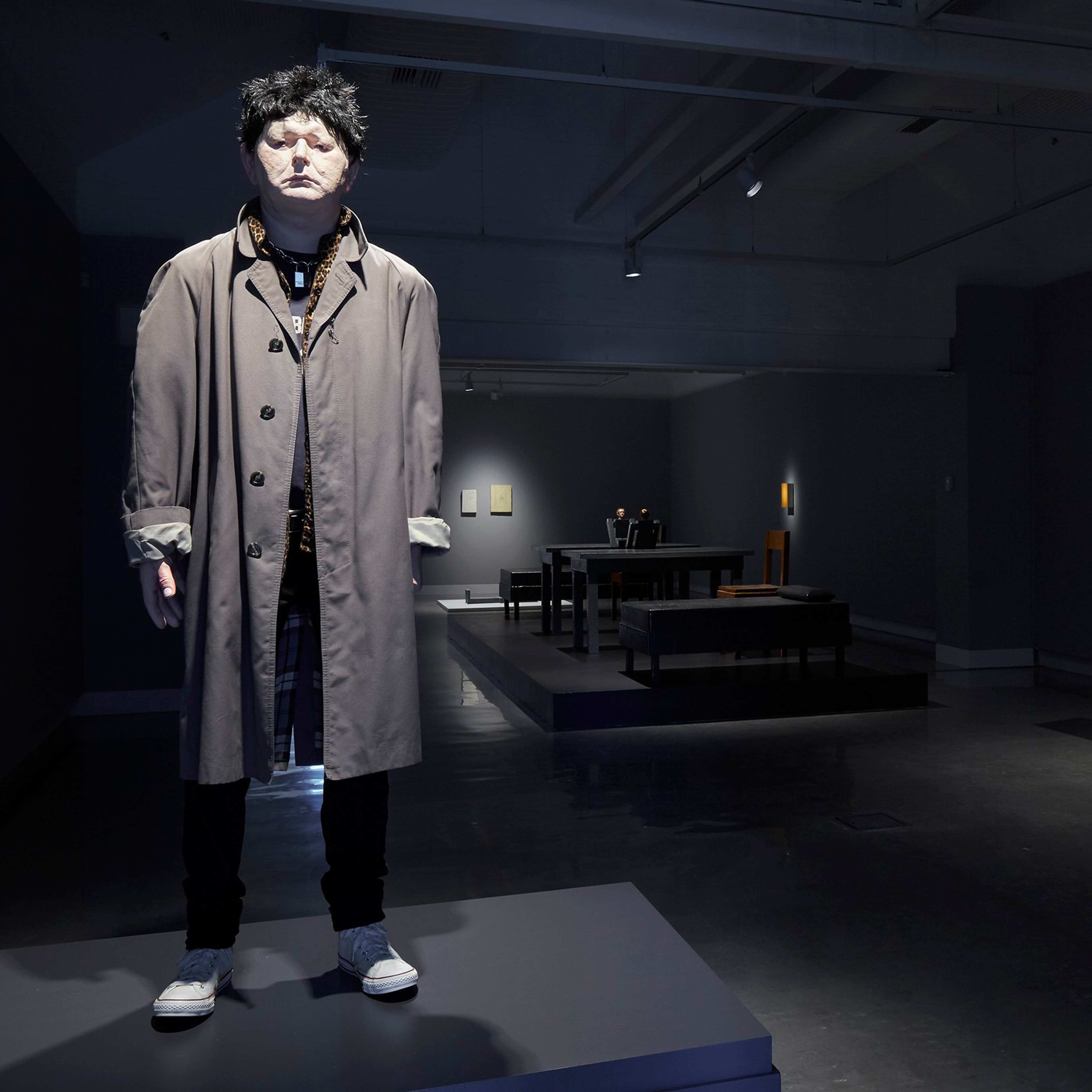 A lifelike mannequin with curly hair, wearing a tan trench coat and sneakers, stands on a pedestal in a dimly lit modern art gallery with various artworks and displays in the background.