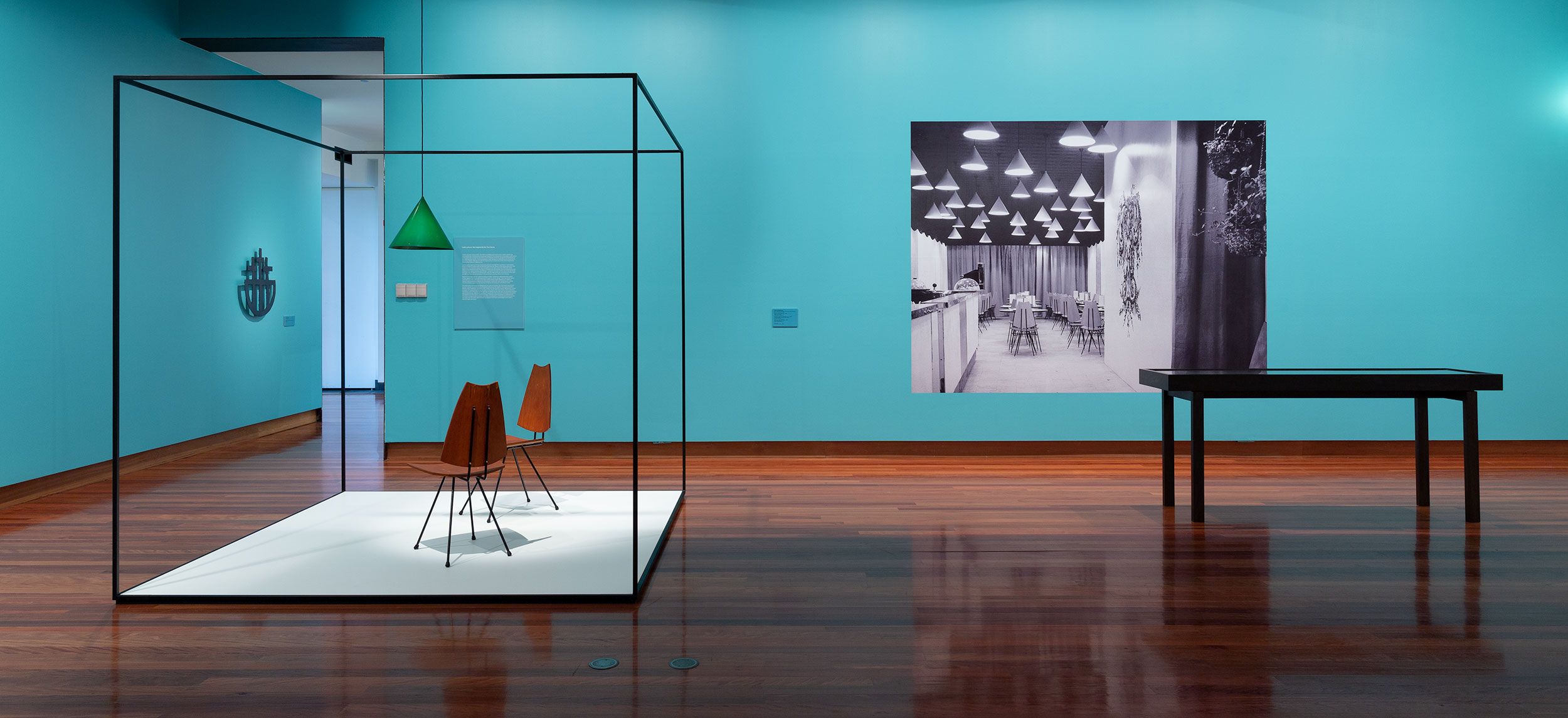A photograph of a gallery space with mid century furniture in the foreground and a black and white photograph of an interior on the aqua-coloured wall