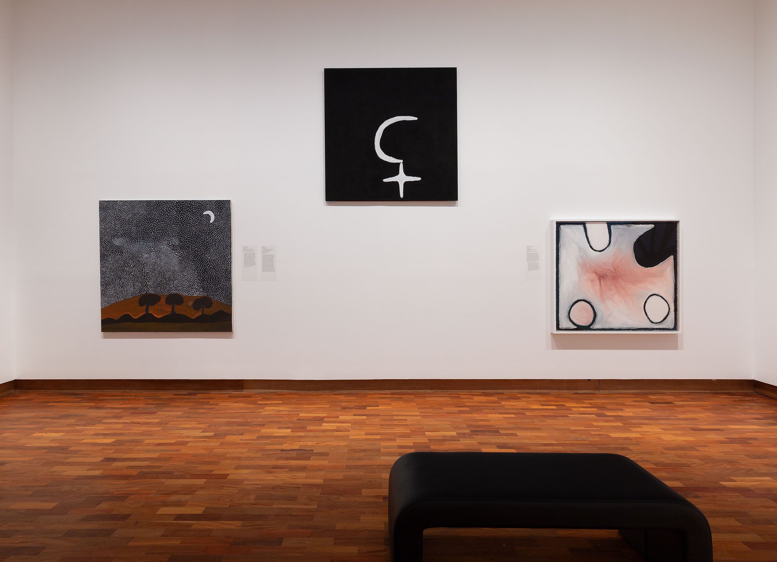 A minimalistInstallation view of 65,000 Years: A Short History of Australian Art, Potter Museum of Art, the University of Melbourne, 2025. Featuring Rusty Peters, Three Nyawana in Yariny Country 2016, Mabel Juli, Garnkiny Ngarranggarni (Moon Dreaming) 2013, and Paddy Bedford, Old Bedford 2005. Collections credit left to right; Hassall Collection, Gadigal Nura (Sydney), National Gallery of Australia, Kamberri (Canberra), Collection of Anthony Scott, Melbourne. Photography by Christian Capurro gallery room with three abstract paintings on a white wall above a wooden floor. A black bench is positioned in front of the artworks.