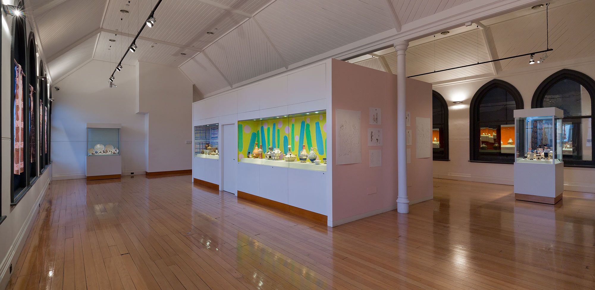 A wide shot of gallery space with multiple vitrines with colourful objects 