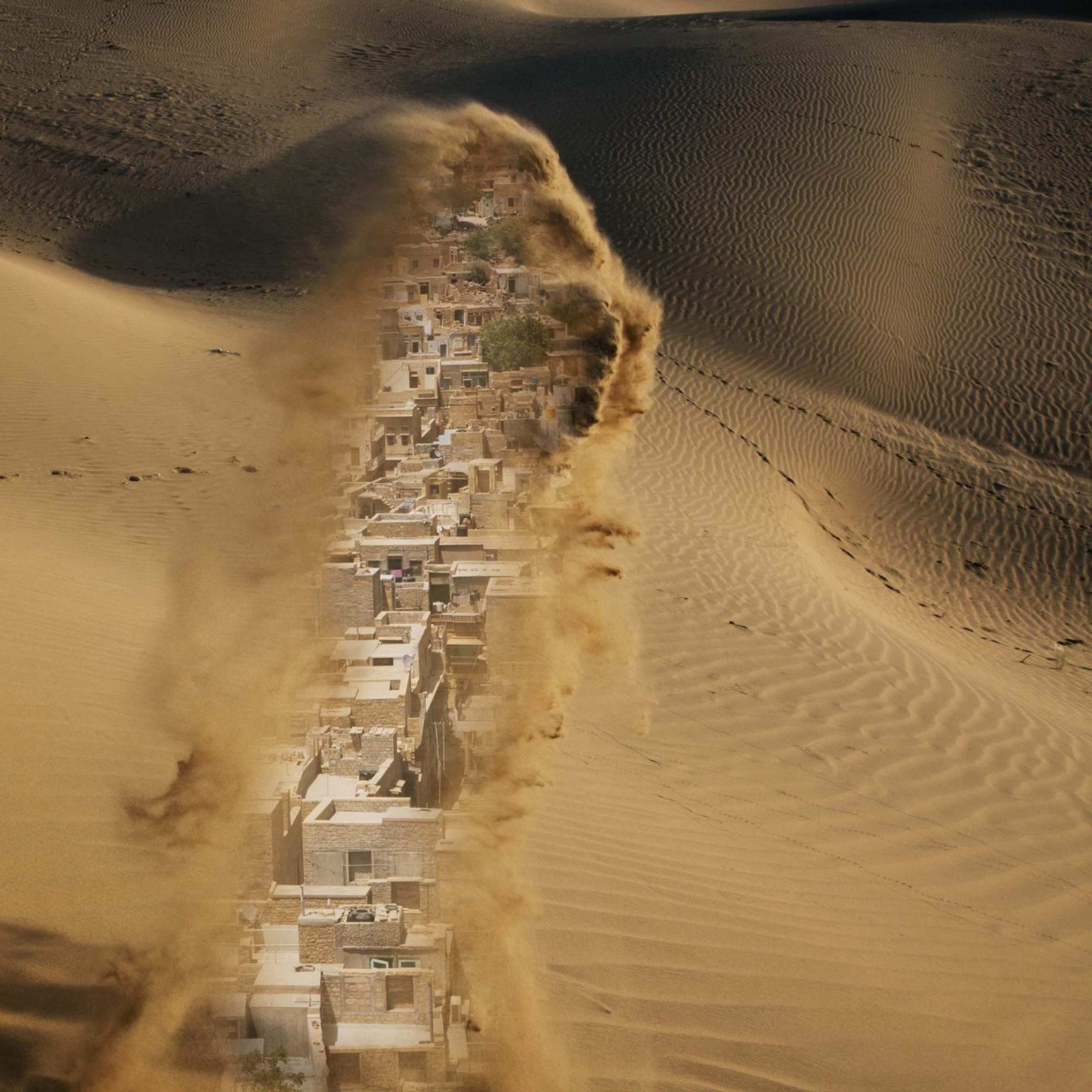 A photograph of a desert sands with a row of buildings partially visible in the moving sand