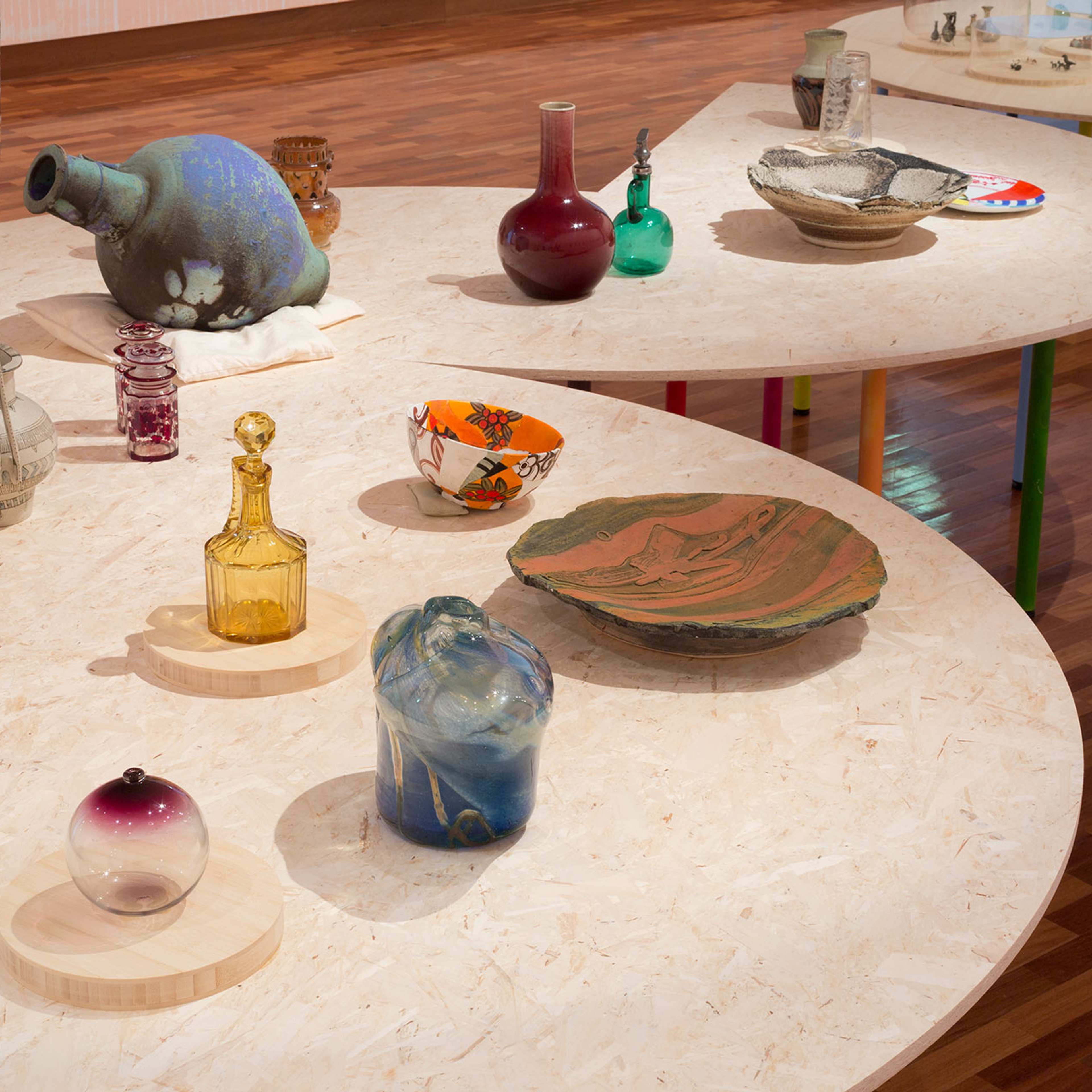 Plates bowls and decorative objects made from various materials are displayed on a plywood table