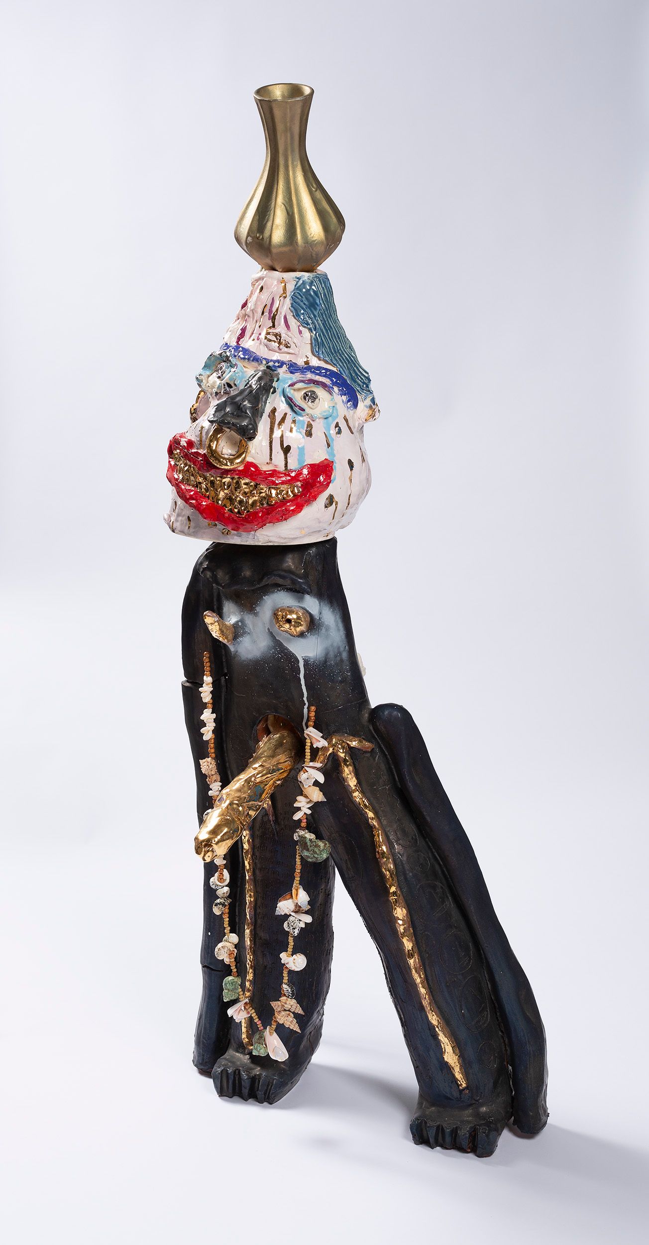 Abstract sculpture featuring a multicolored face with large red lips, golden accents, and a textured surface. The figure has long black legs, an elongated torso adorned with hanging beads, and a gold vase-like object on top.