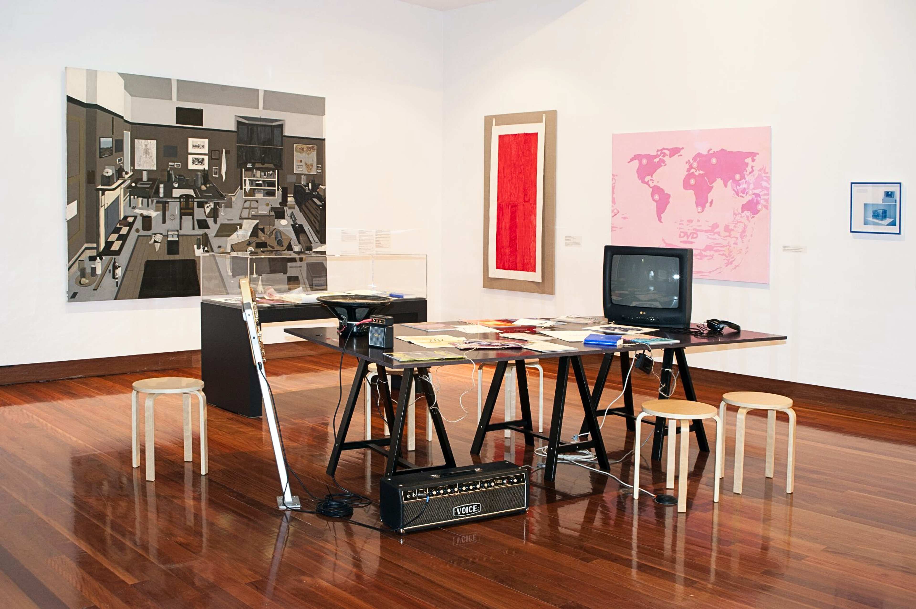 A photograph of an exhibition space with a large table holding a tv, and various publications in the centre, an guitar amplifier on the floor next to the table, and large format drawings and photographs on the walls around
