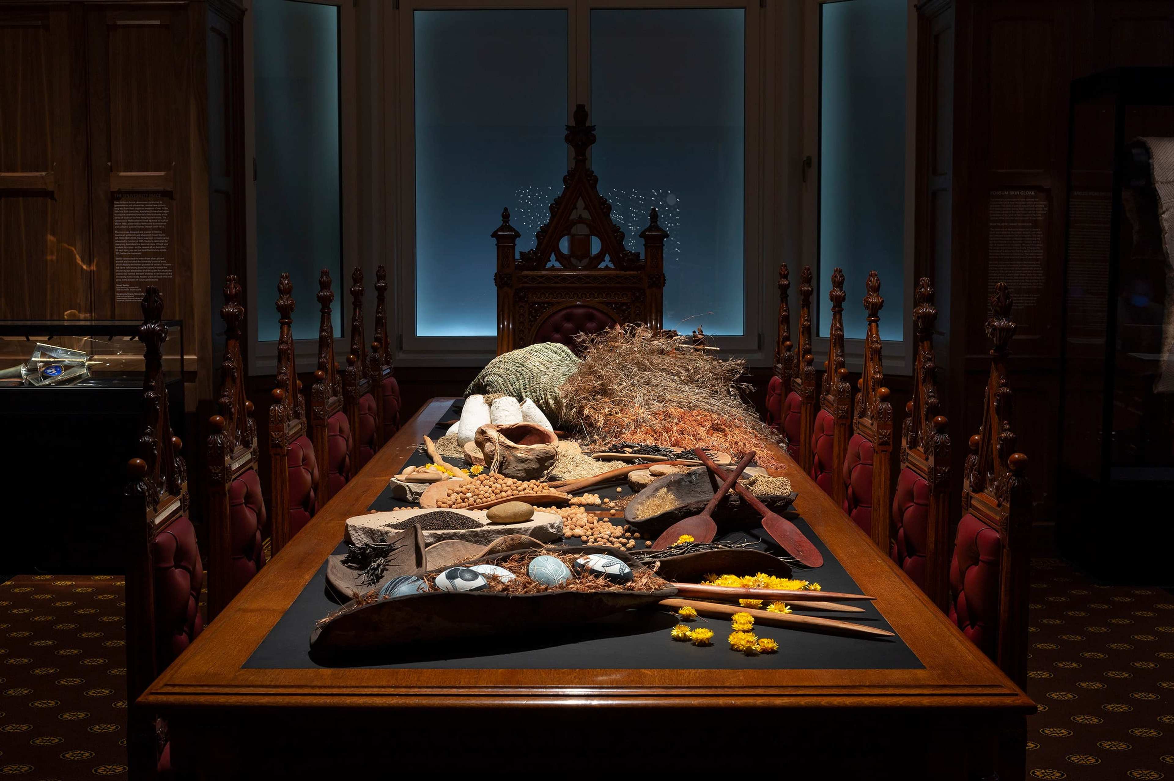 A long wooden table with ornate chairs displays various natural and cultural objects, including shells, stones, woven items, and wooden tools, in a dimly lit room with a large, carved chair at the end.
