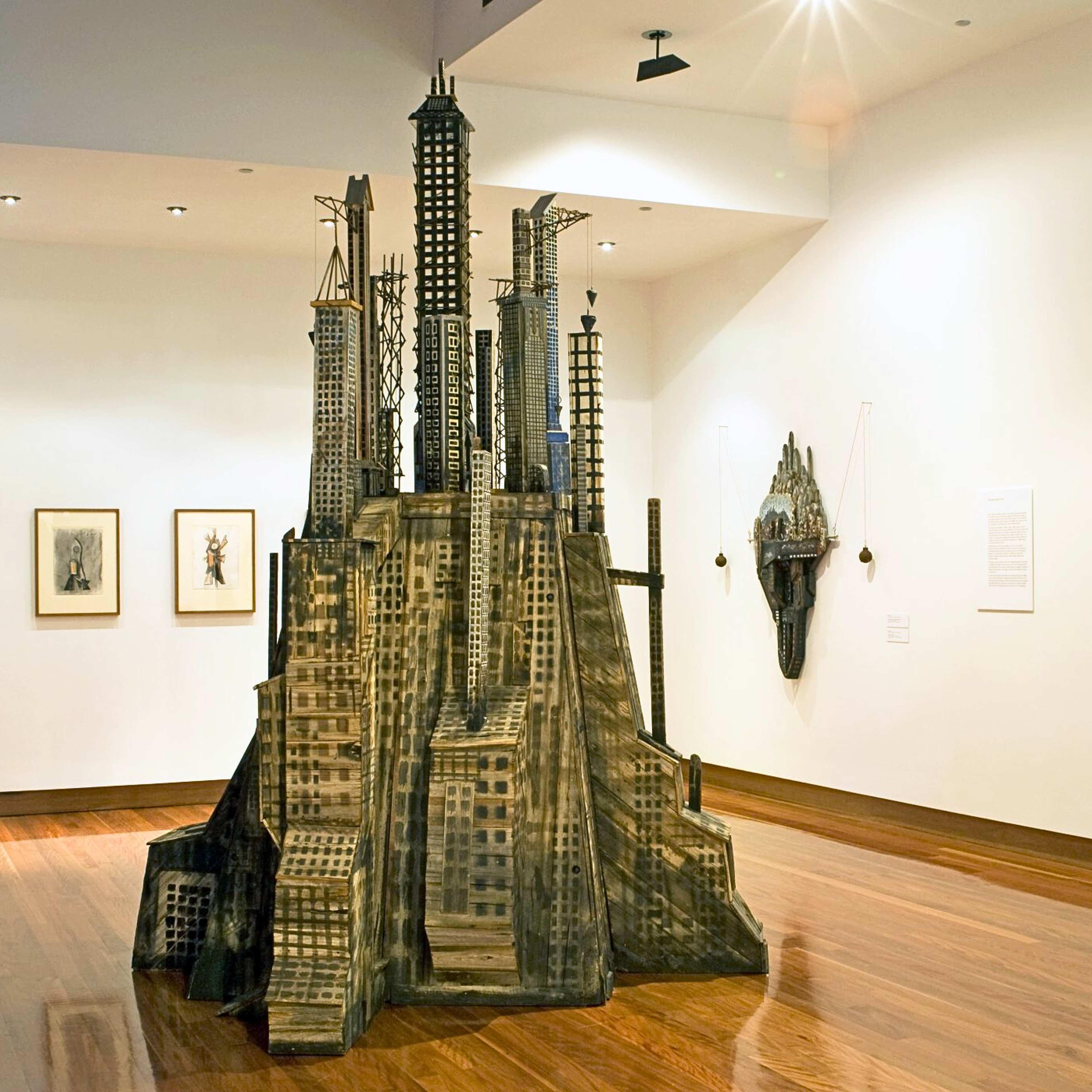 A photograph of a gallery featuring large-scale sculpture reminiscent of a coup of skyscrapers crowded over a hill