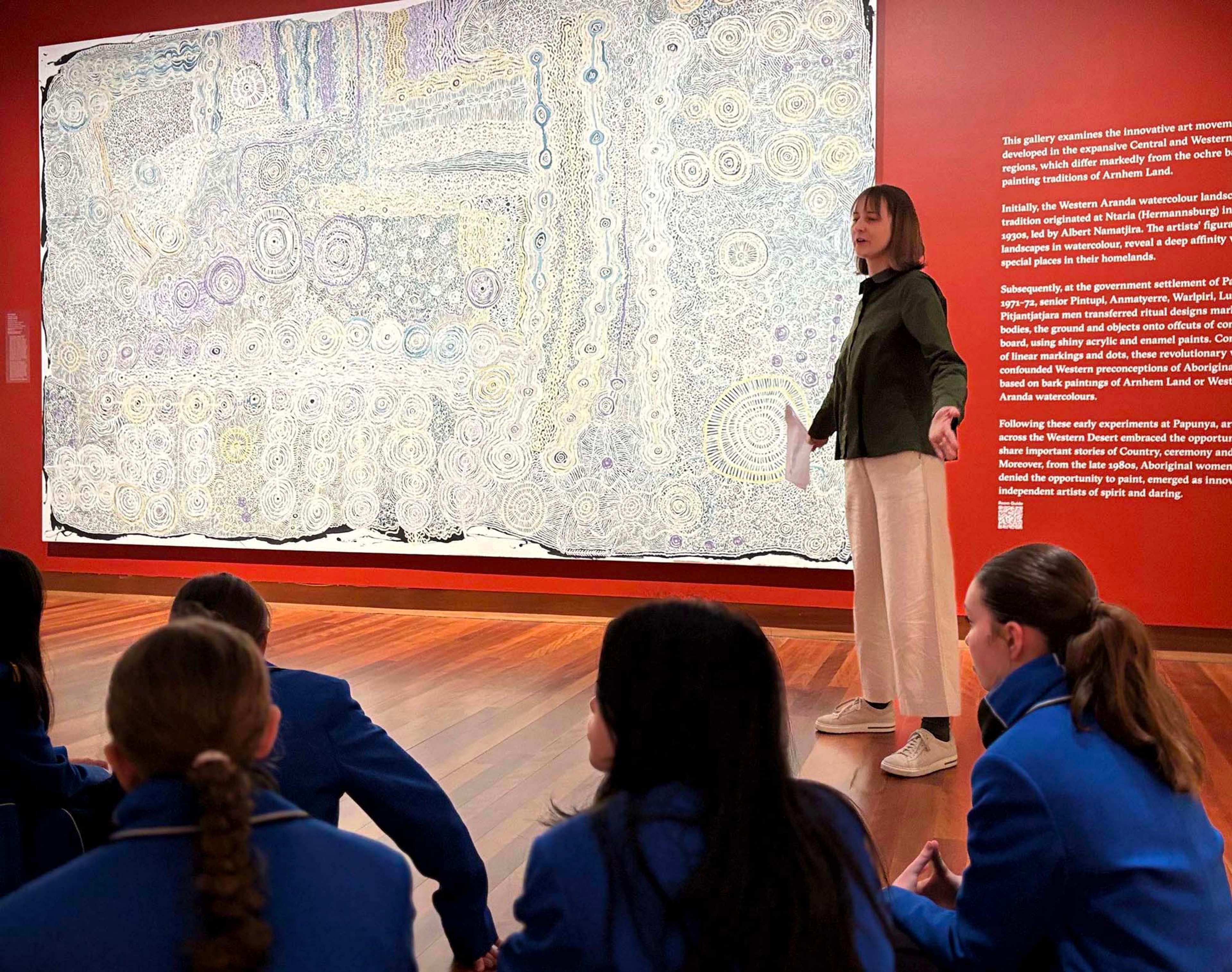 A person speaks to a group of students in blue uniforms seated on the floor in front of a large, detailed abstract artwork displayed on a red gallery wall.