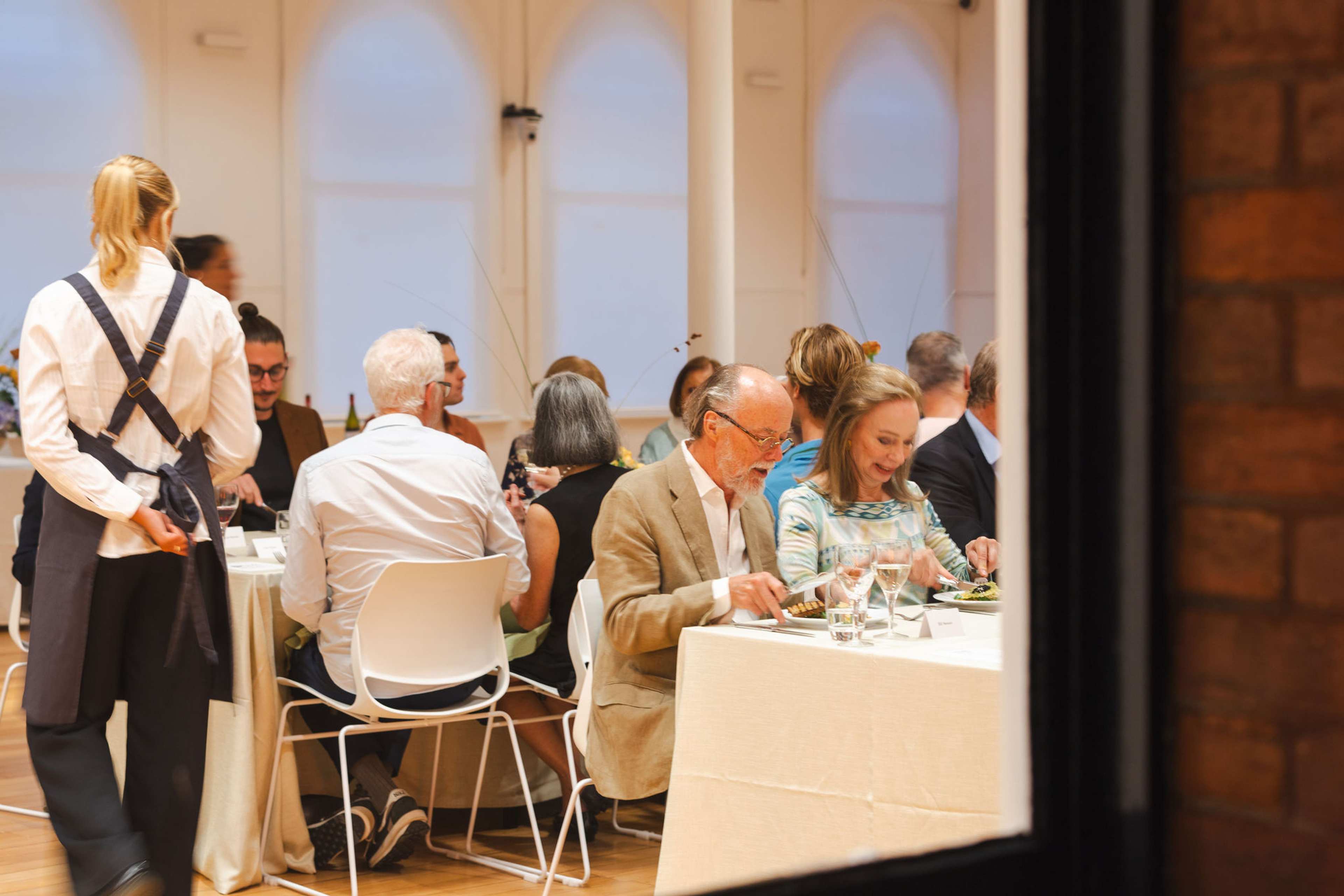 A formal dining setup with white chairs and tables decorated with blue flowers with people eating and waitress serving