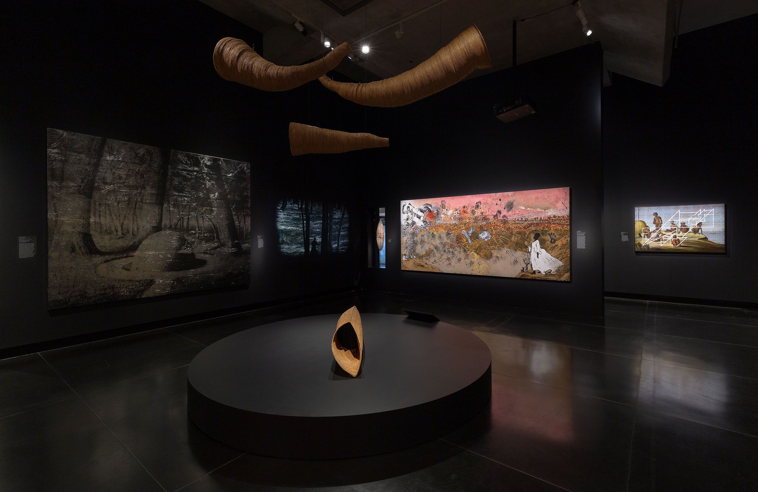 A dark gallery displays hanging sculptural eel traps, a woven sculptural eel trap on a round platform, and three large paintings featuring abstract and natural themes, all under focused lighting.