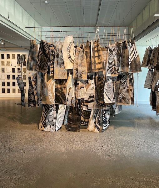 Textile panels with abstract black, brown, and white patterns hang from wires in an art gallery with concrete floors and bright natural light. Other artwork is visible in the background.