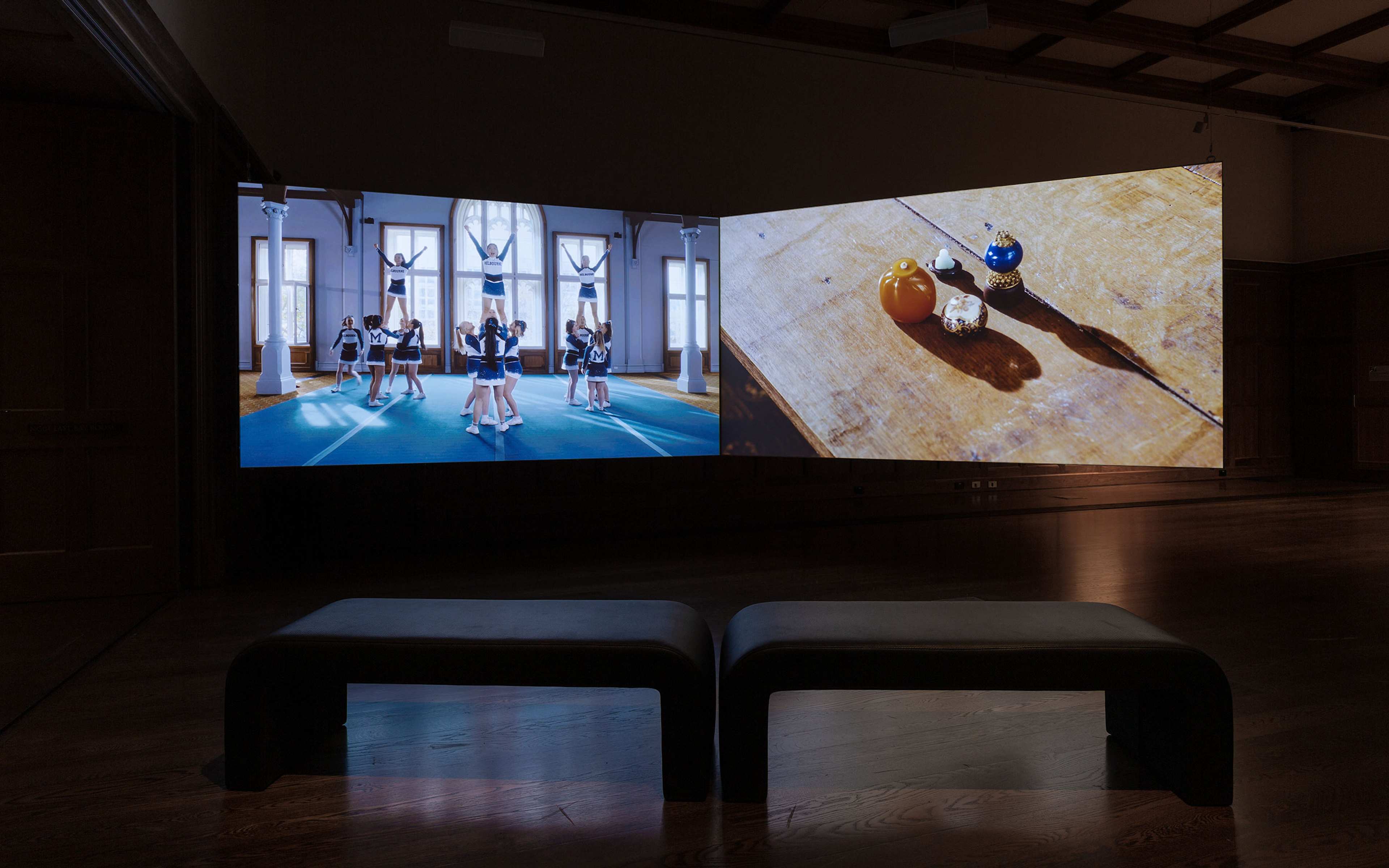 Two screens in a gallery show a video of cheeleaders with arms in air and a video of doorknobs on wooden floor.