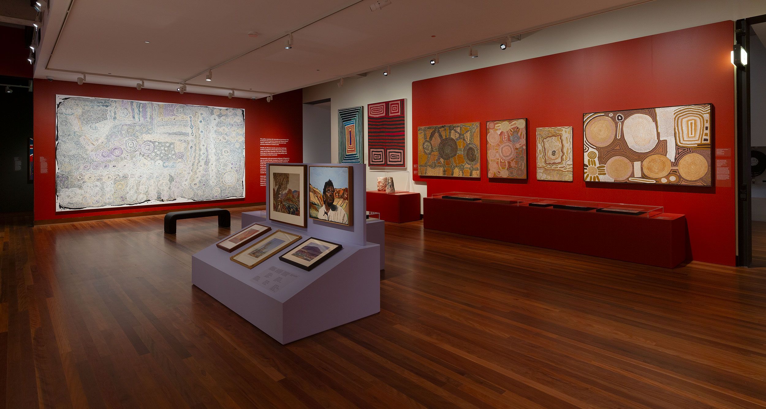 A gallery with red walls displays Indigenous Australian paintings in earthy tones. Framed artworks are mounted on walls and stands, and a bench sits in front of a large, detailed painting. The floor is wooden.