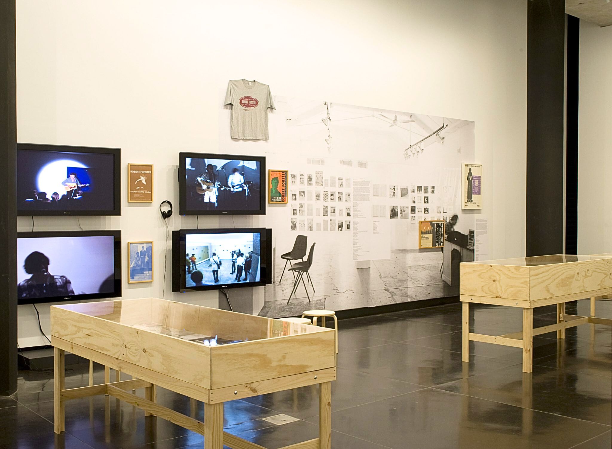 A photograph of a gallery with multiple vitrines, tv screens, a t-shirt and a large paste up on the wall