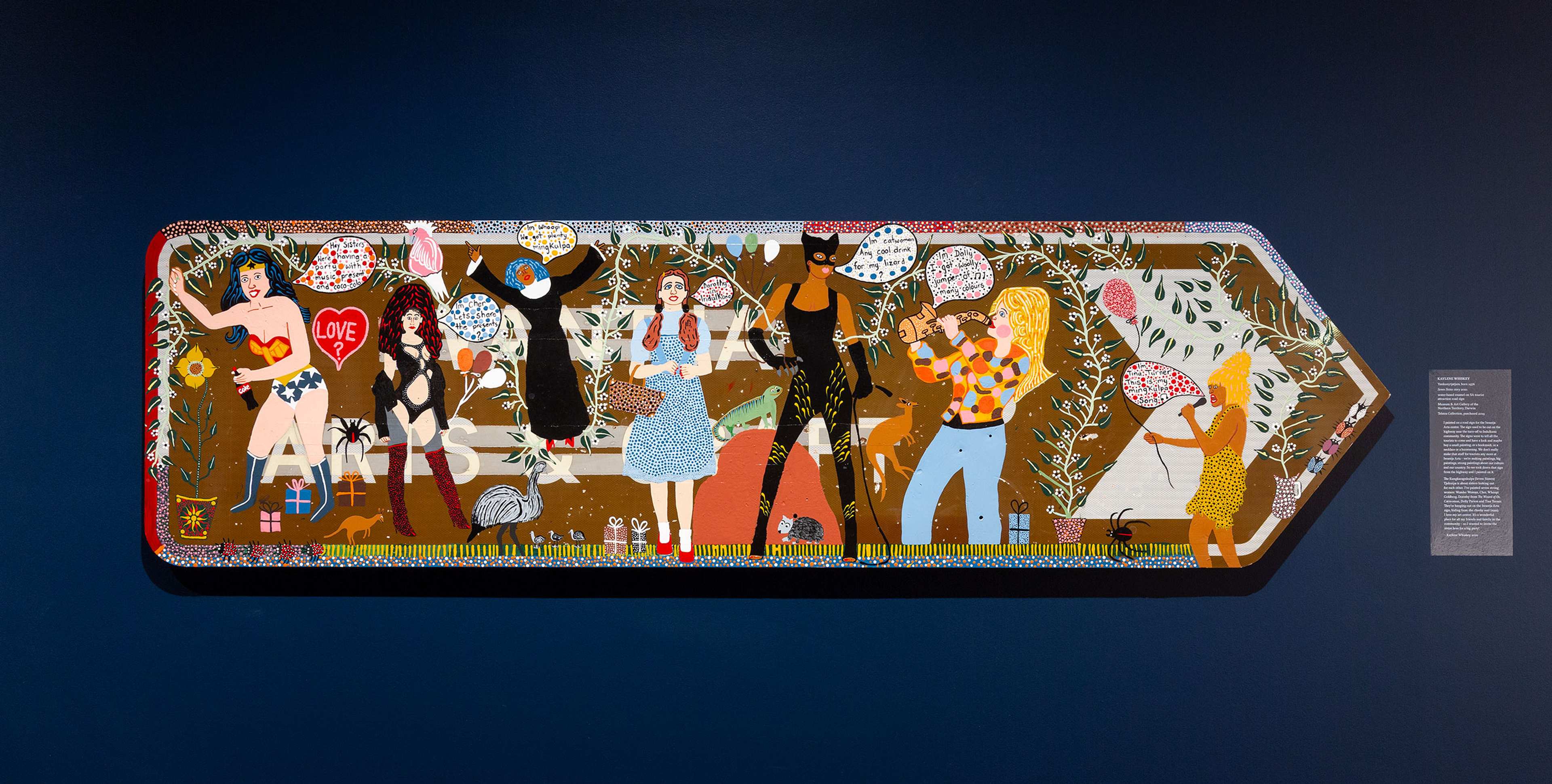 Colounstallation view of 65,000 Years: A Short History of Australian Art, Potter Museum of Art, the University of Melbourne, 2025. Featuring Kaylene Whiskey (Yankunytjatjara, born 1976), Seven Sistas story 2021. Museum & Art Gallery of the Northern Territory, Darwin. Photography by Christian Capurro.rful artwork shaped like a large arrow features diverse cartoon-like figures, speech bubbles, and symbolic objects on a dark blue wall, illustrating various interactions and narratives in a vibrant, playful style.