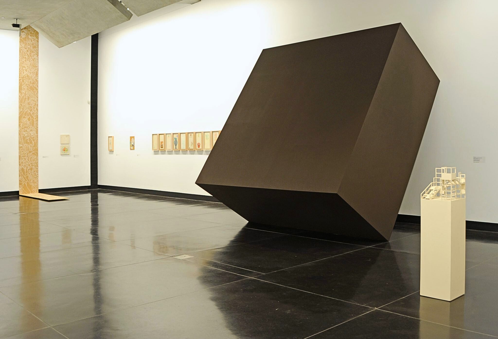 A photograph of a gallery with various works including a giant black cube leaning against the wall and a white architectural model on the plinths in front of it