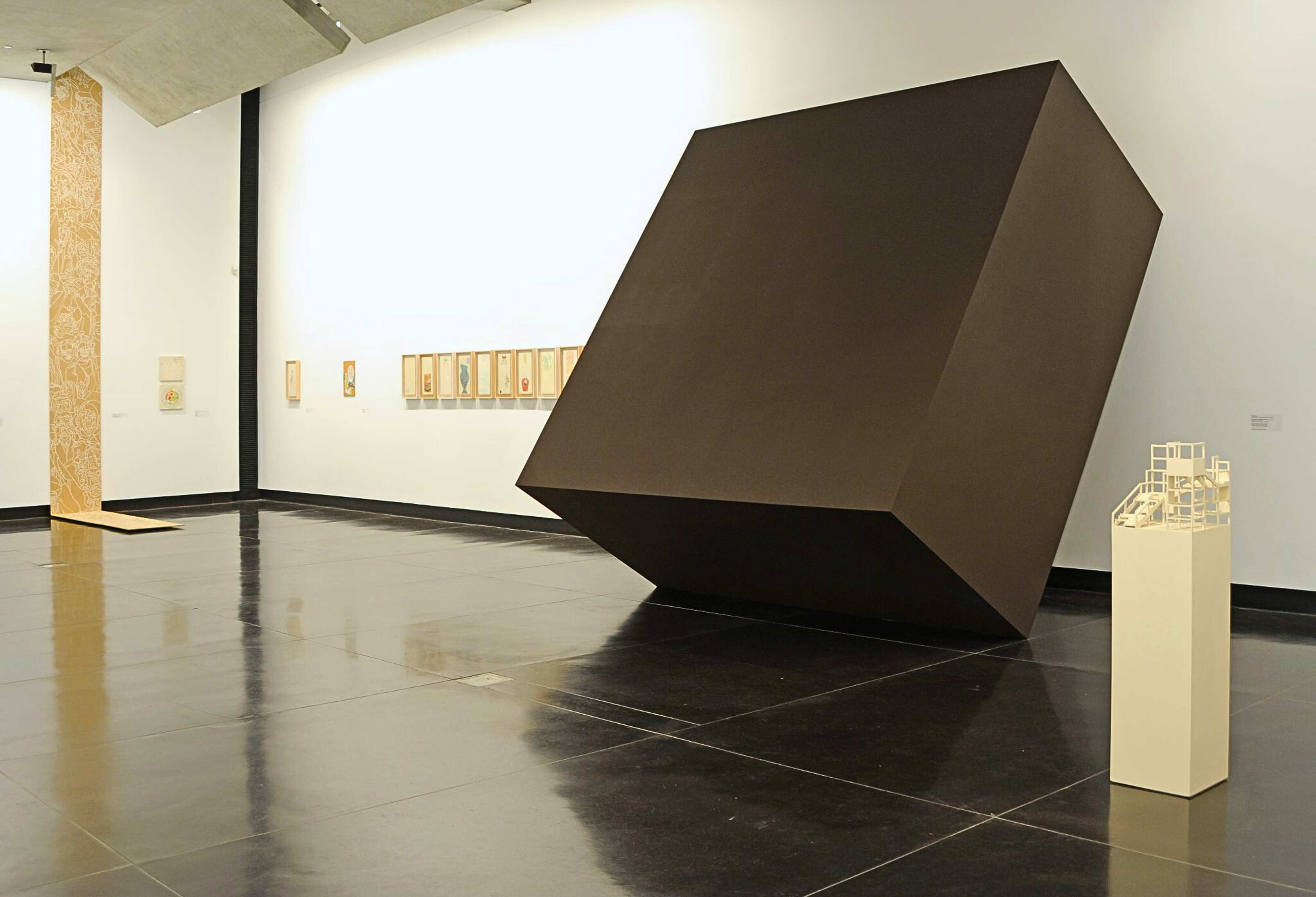 A photograph of a gallery with various works including a giant black cube leaning against the wall and a white architectural model on the plinths in front of it